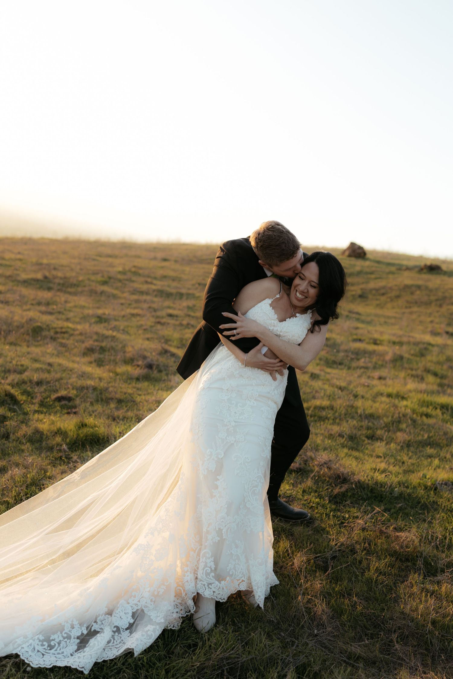 A couple in wedding attire share a romantic dip pose together in a grassy field at sunset.
