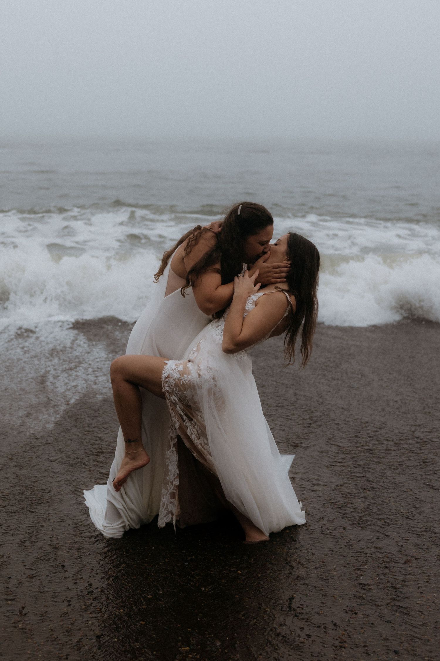 A couple in white outfits share intimate moments on a dark sandy beach with crashing waves in the background.
