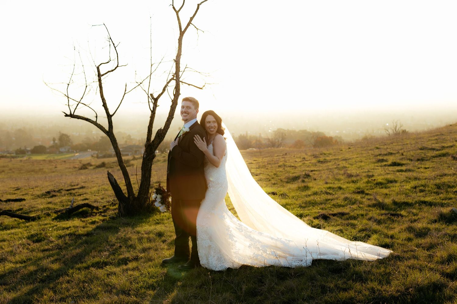 A romantic wedding couple stands together in a misty field at sunset with a bare tree in the background.