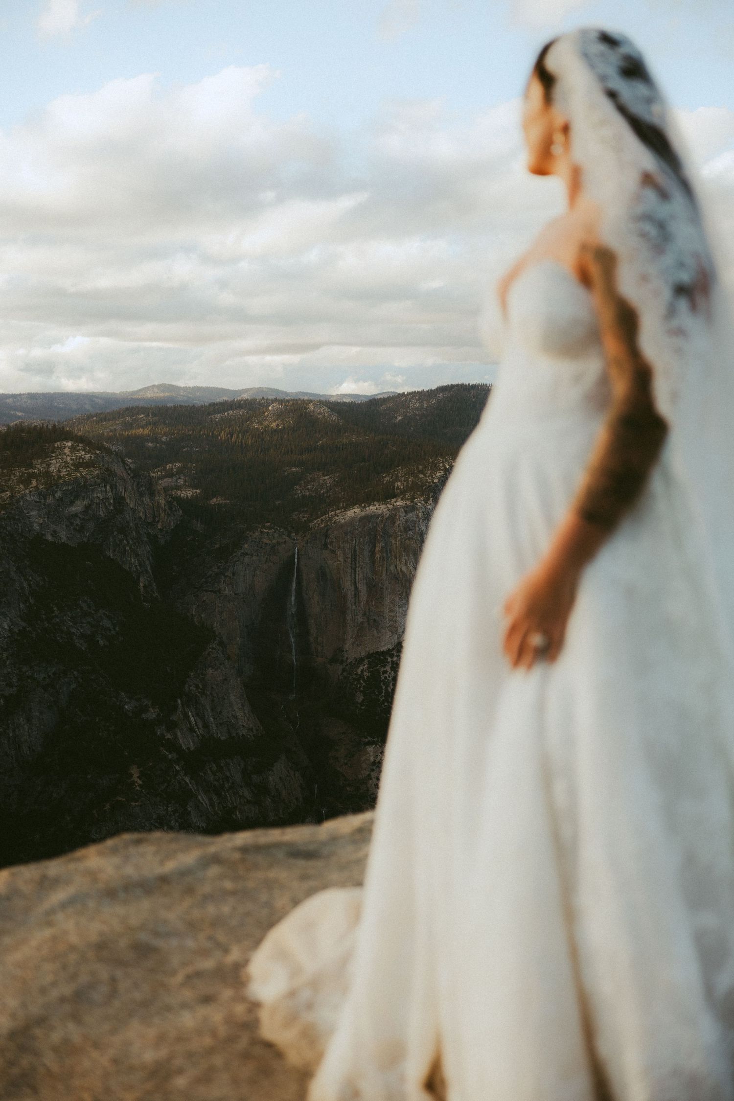 Bride in white gown and veil stands on rocky overlook with stunning mountain valley views at Yosemite.