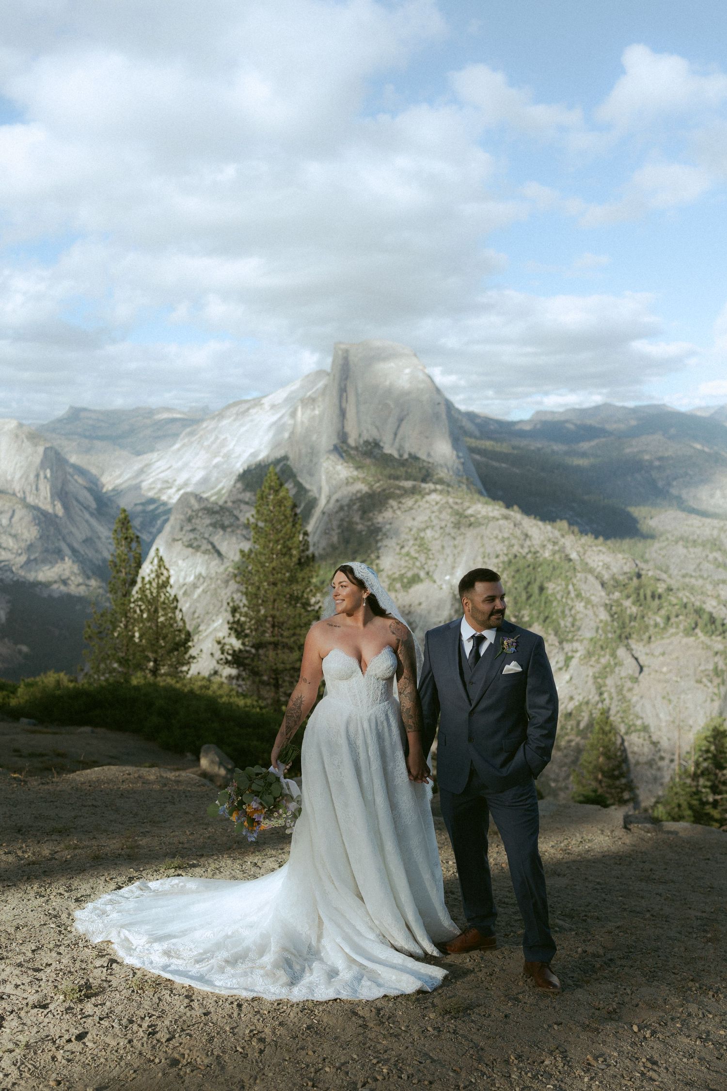 Bride and groom hold hands at Glacier Point with Half Dome and Yosemite Valley in the background.