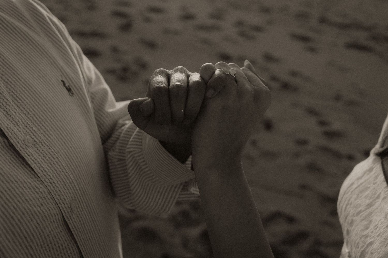 Black and white photo of a couple holding hands on a sandy beach, wearing a wedding ring.