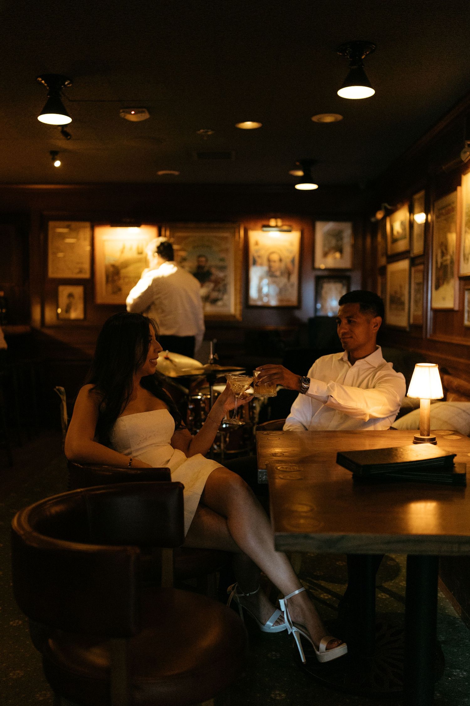 Couple sharing drinks at a dimly lit bar, woman in white dress and man in white shirt in romantic setting.