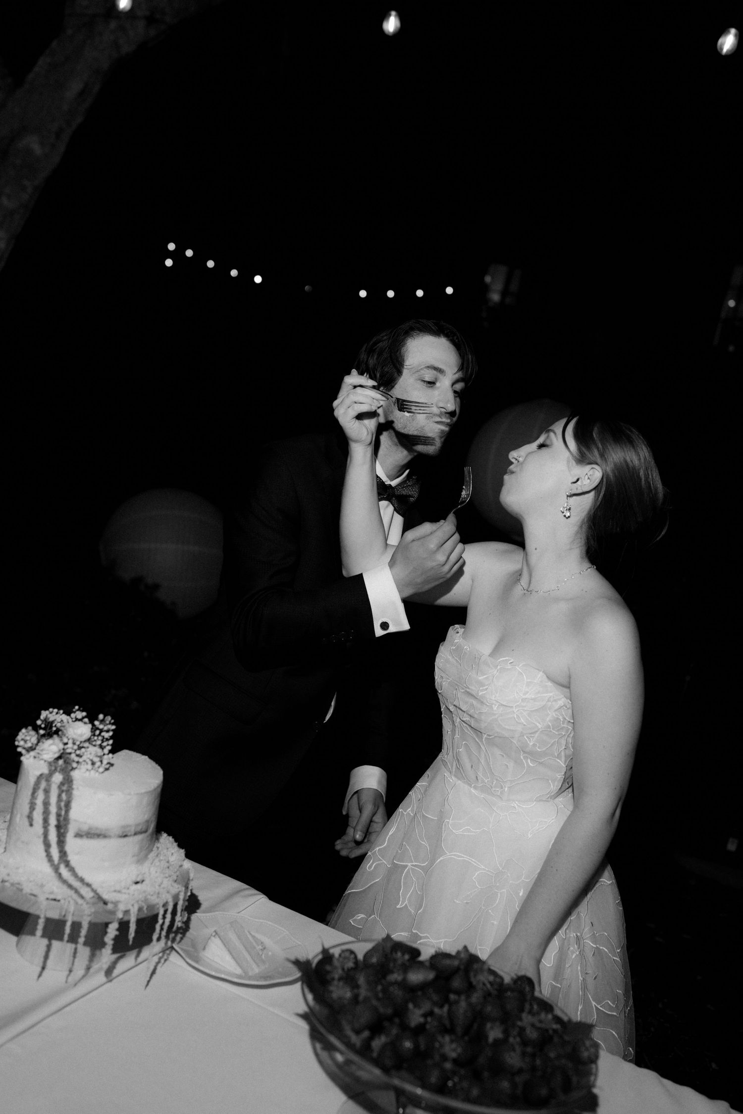 Black and white photo of bride and groom sharing wedding cake at outdoor evening reception with string lights.