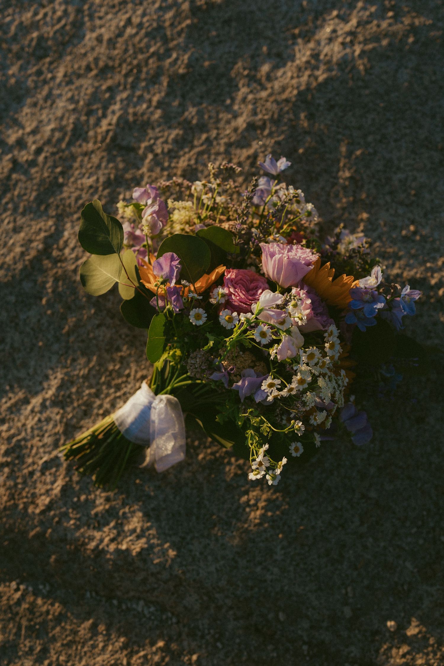 Colorful wildflower wedding bouquet with pink, purple, and white blooms resting on sandy ground in warm sunlight.
