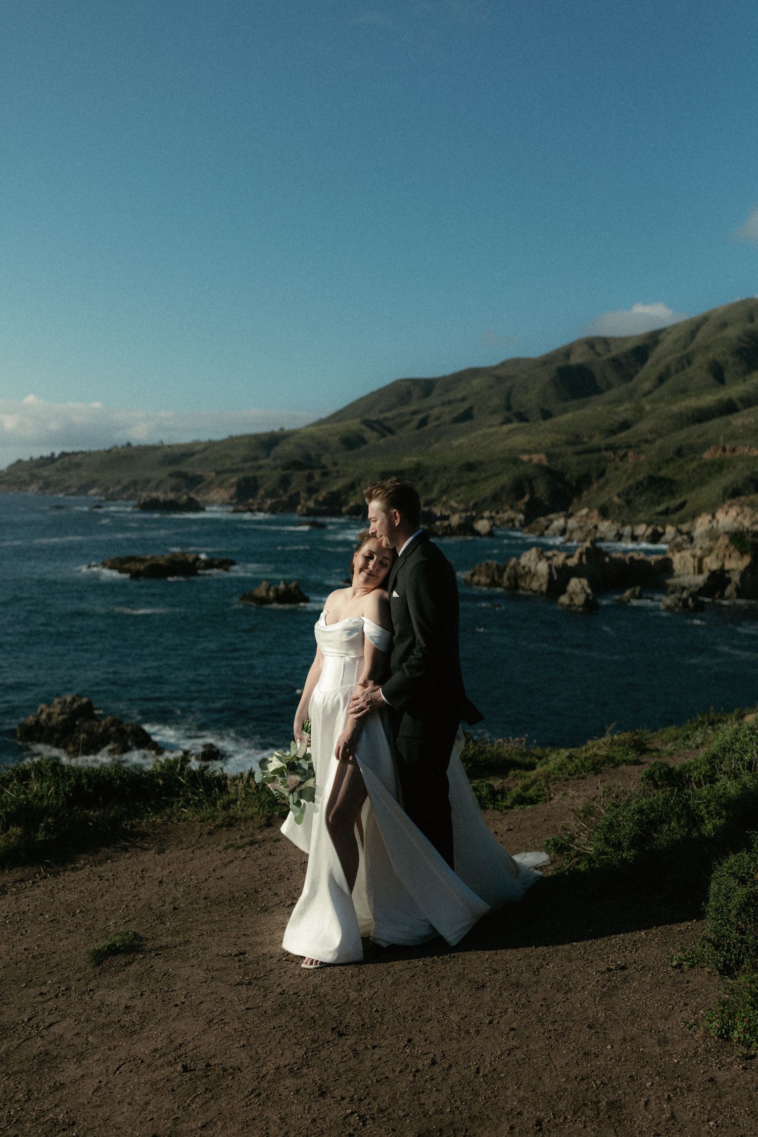 A bride and groom embrace on a coastal cliffside with stunning ocean views and green hills in Big Sur, California.