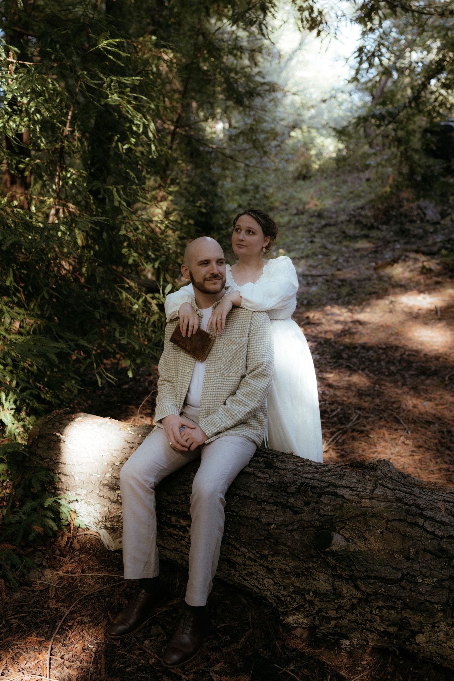 A couple in light colored clothing sits intimately on a fallen tree trunk in a sunlit forest.