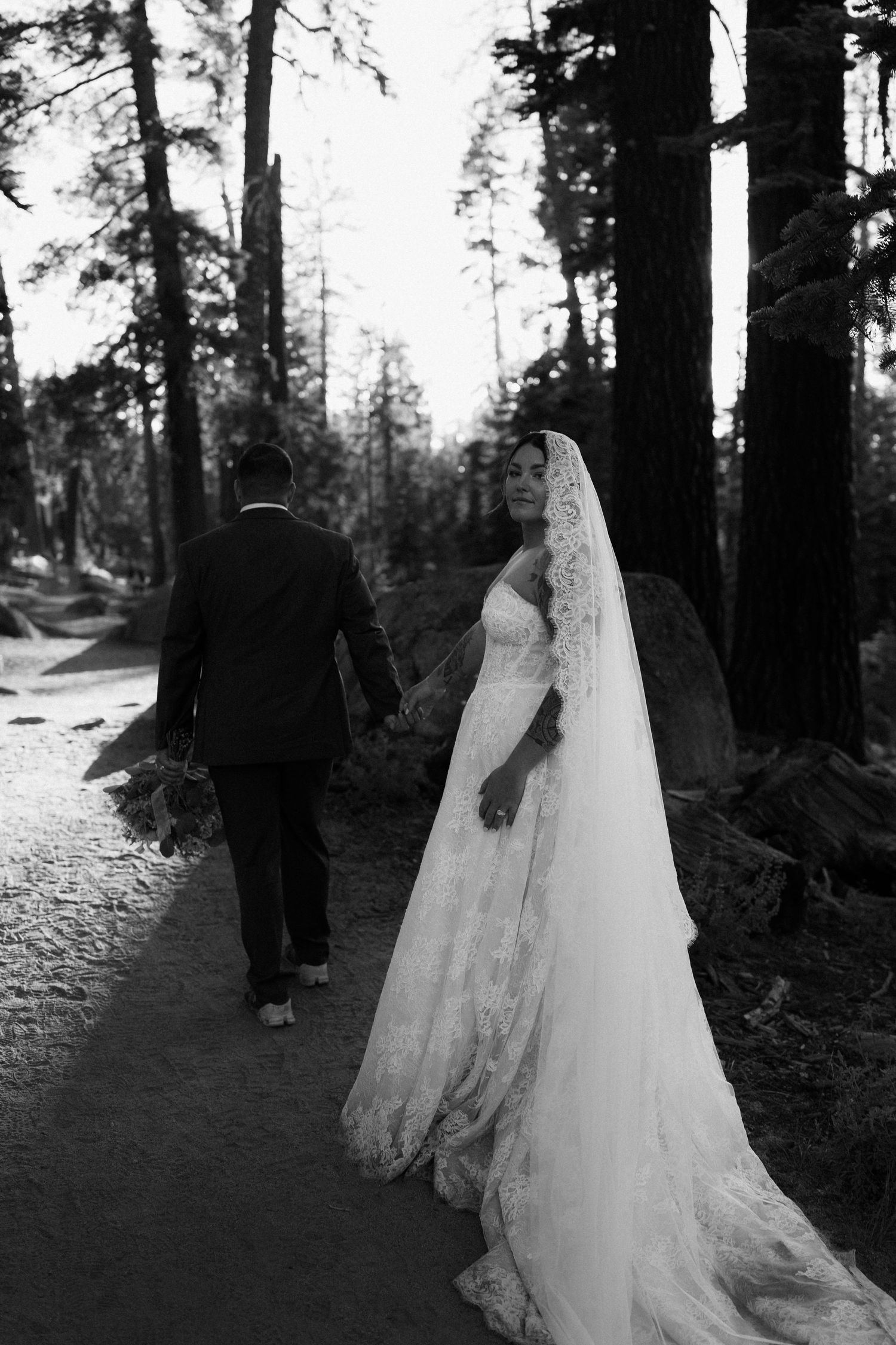 Bride and groom walk hand in hand through a forest path, bride wearing a lace gown and long veil, black and white photo.