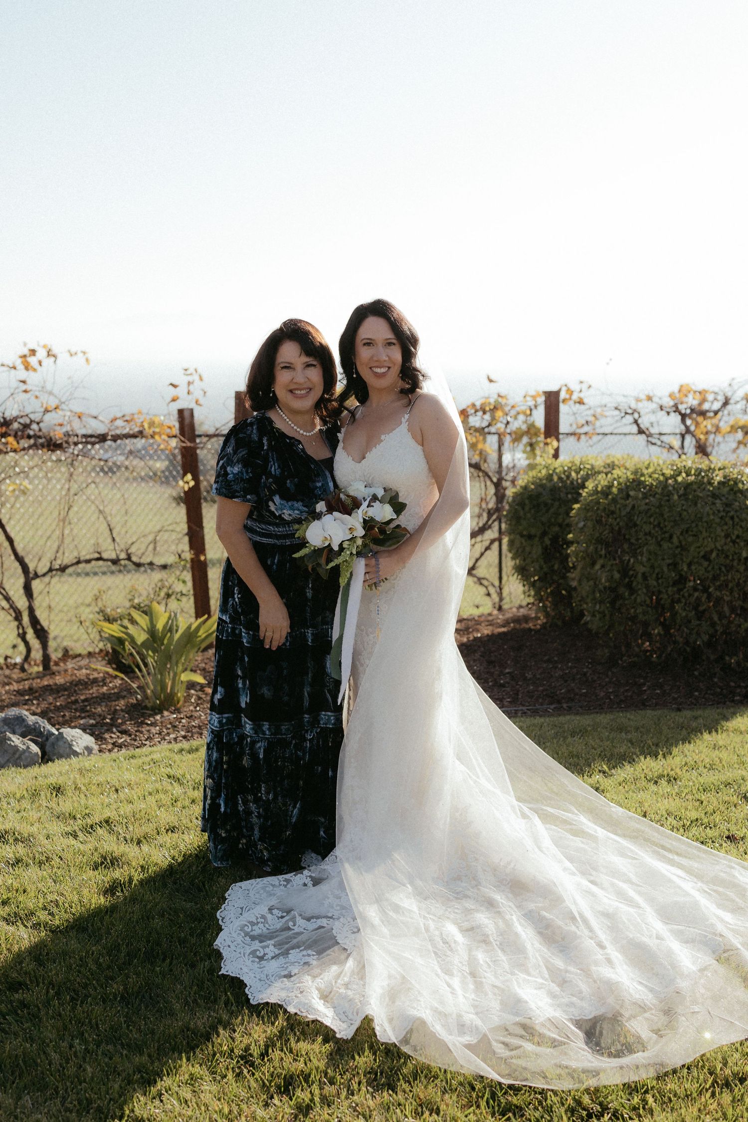Two people pose together outdoors in wedding attire with one wearing a flowing white dress with long train.