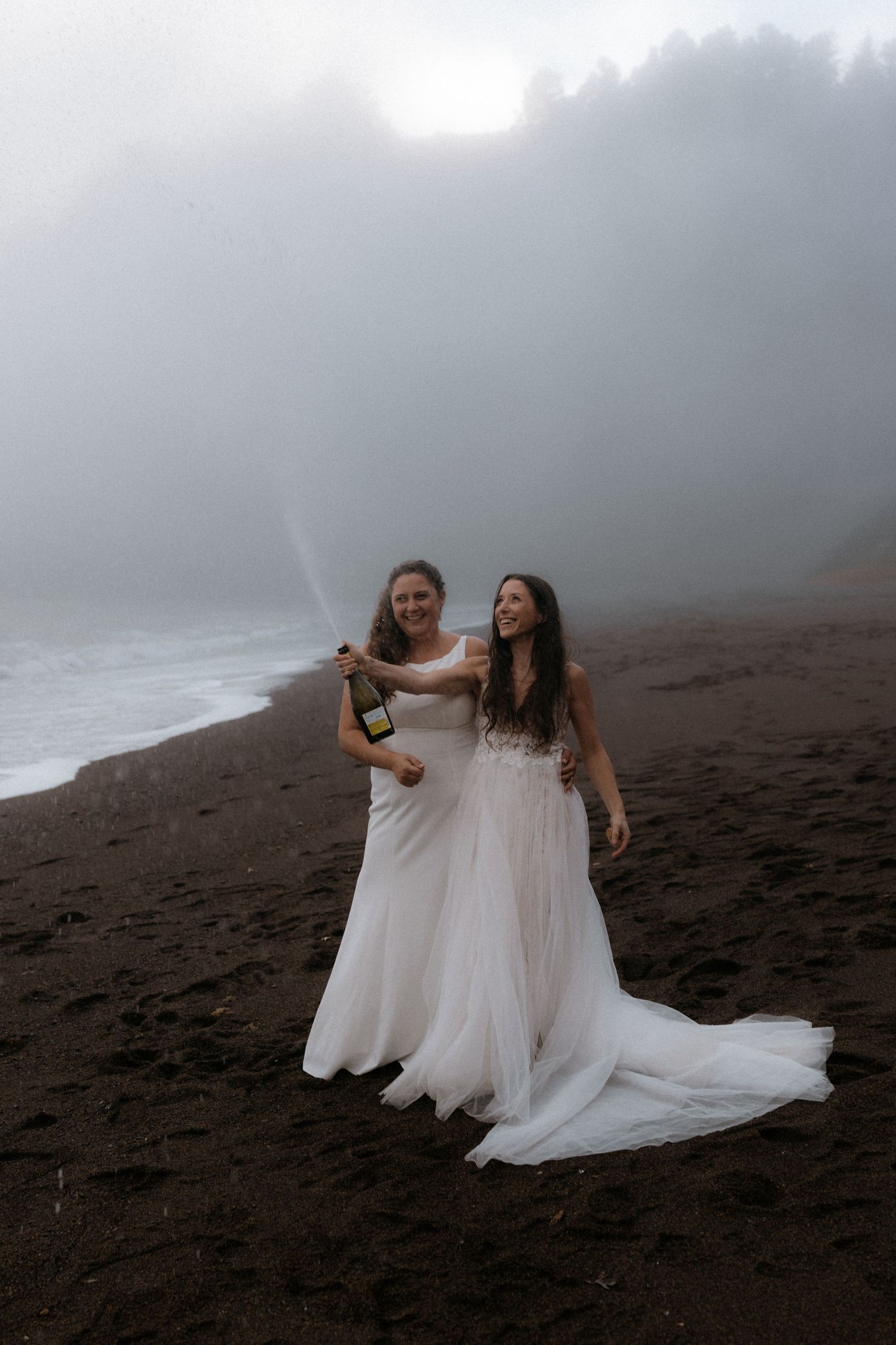 Two people in flowing white dresses pose together on a misty volcanic beach during a photoshoot.