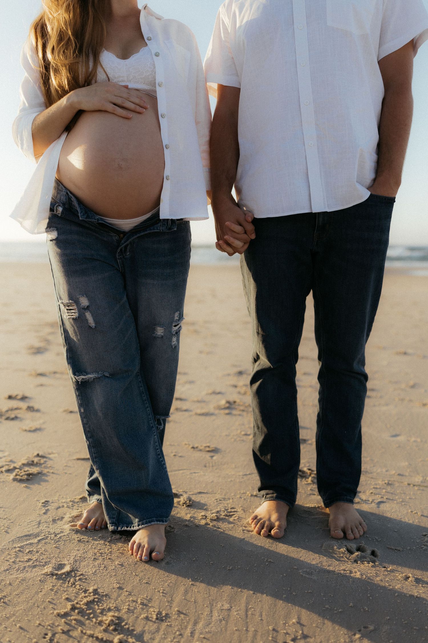 Pregnant woman in ripped jeans holds her baby bump while holding hands with partner on sandy beach.