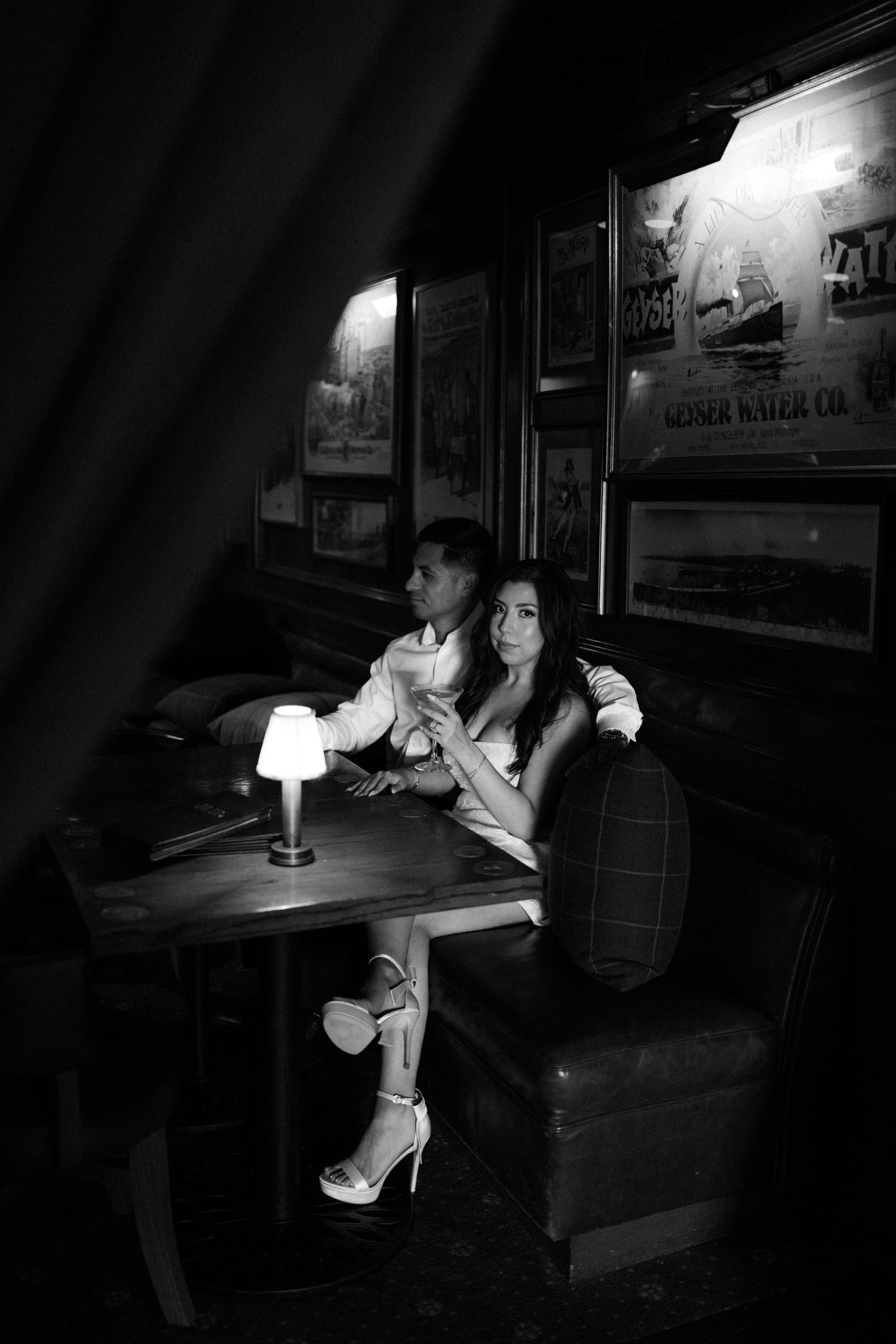 Black and white photo of a couple sitting intimately at a dimly lit table in a vintage bar with framed artwork.