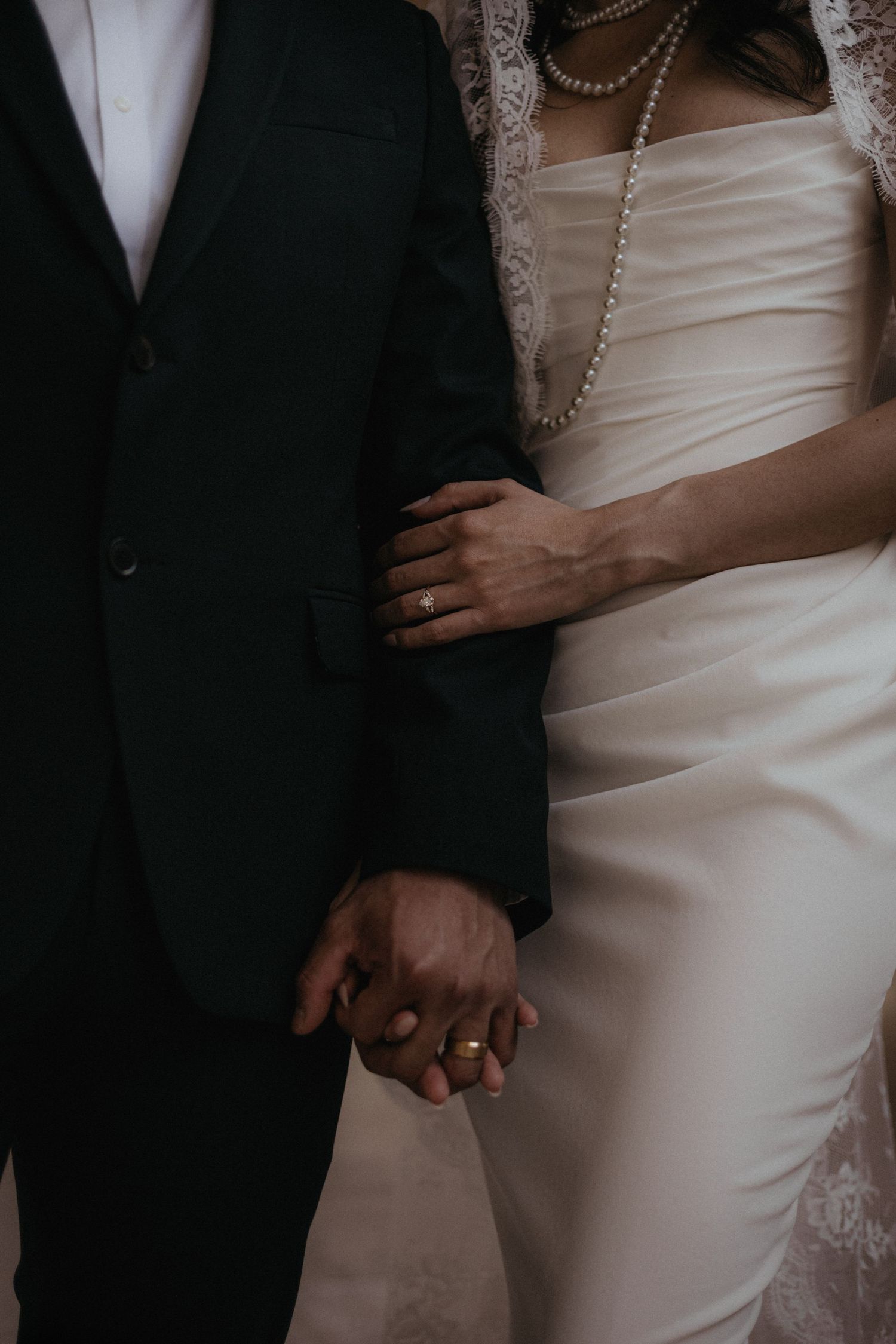 Bride and groom holding hands, wearing wedding rings, in elegant white gown with pearls and dark green suit.
