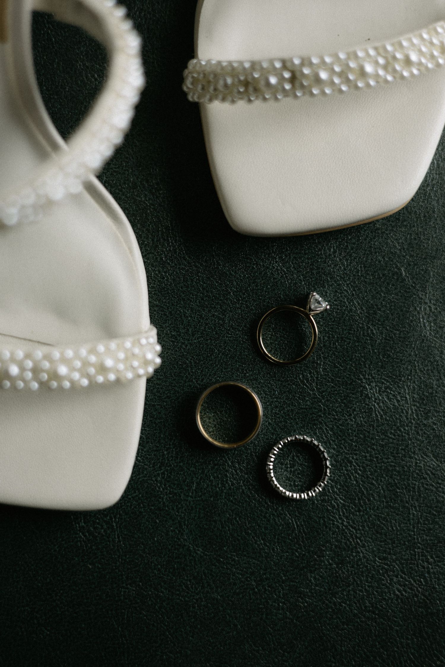 White pearl-embellished bridal heels displayed with three wedding rings on a dark green leather surface.