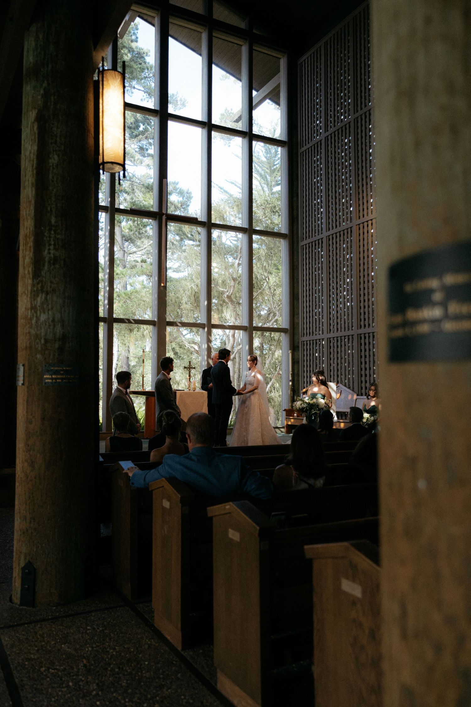 Intimate chapel wedding ceremony with couple exchanging vows before tall windows with forest views.