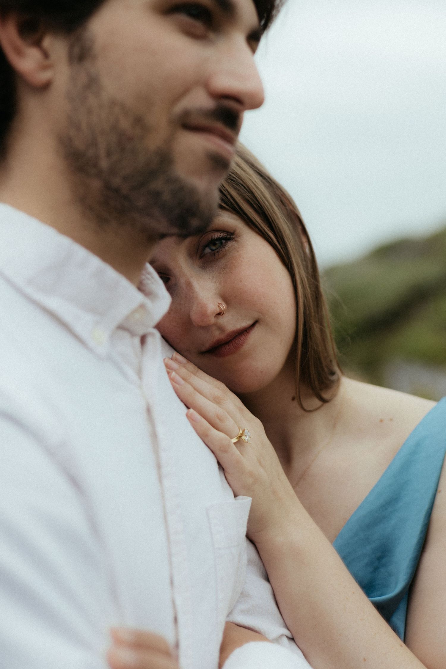 A couple shares an intimate moment, woman in blue dress rests her cheek on mans chest showing gold engagement ring.