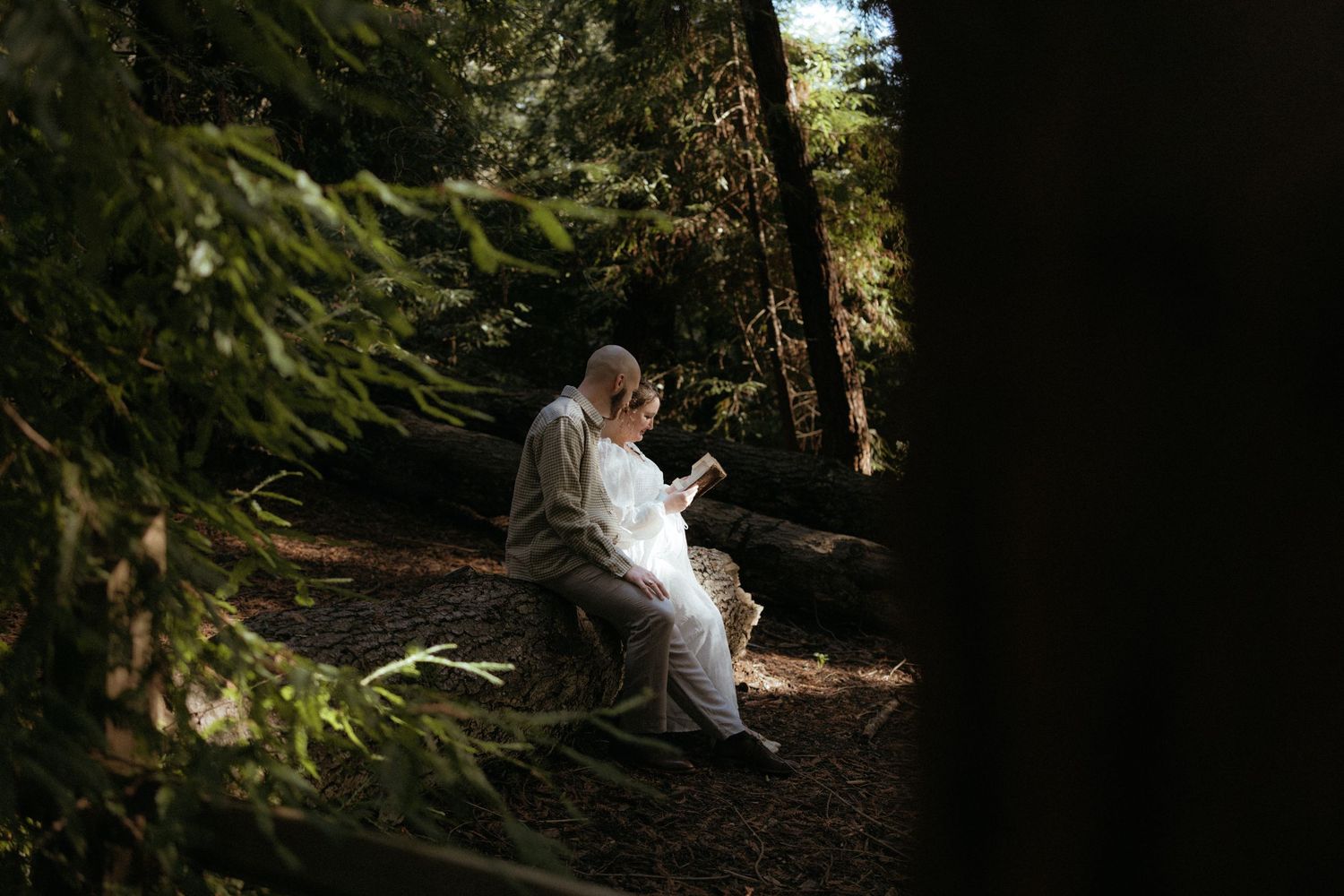 A couple in white wedding attire embraces while walking through a sunlit forest path.