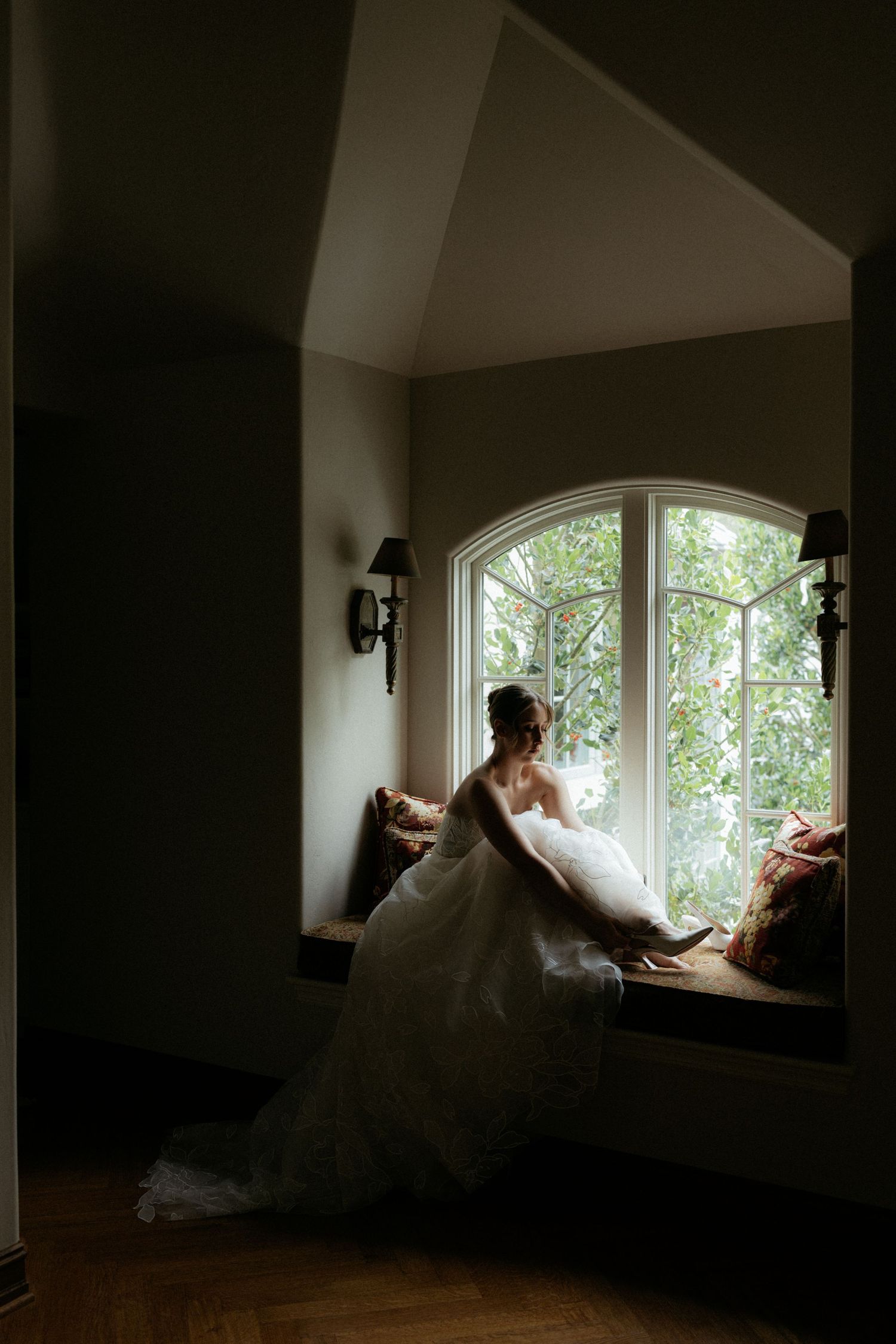 Bride in white gown sitting on window seat gazing outside through arched window in softly lit room.