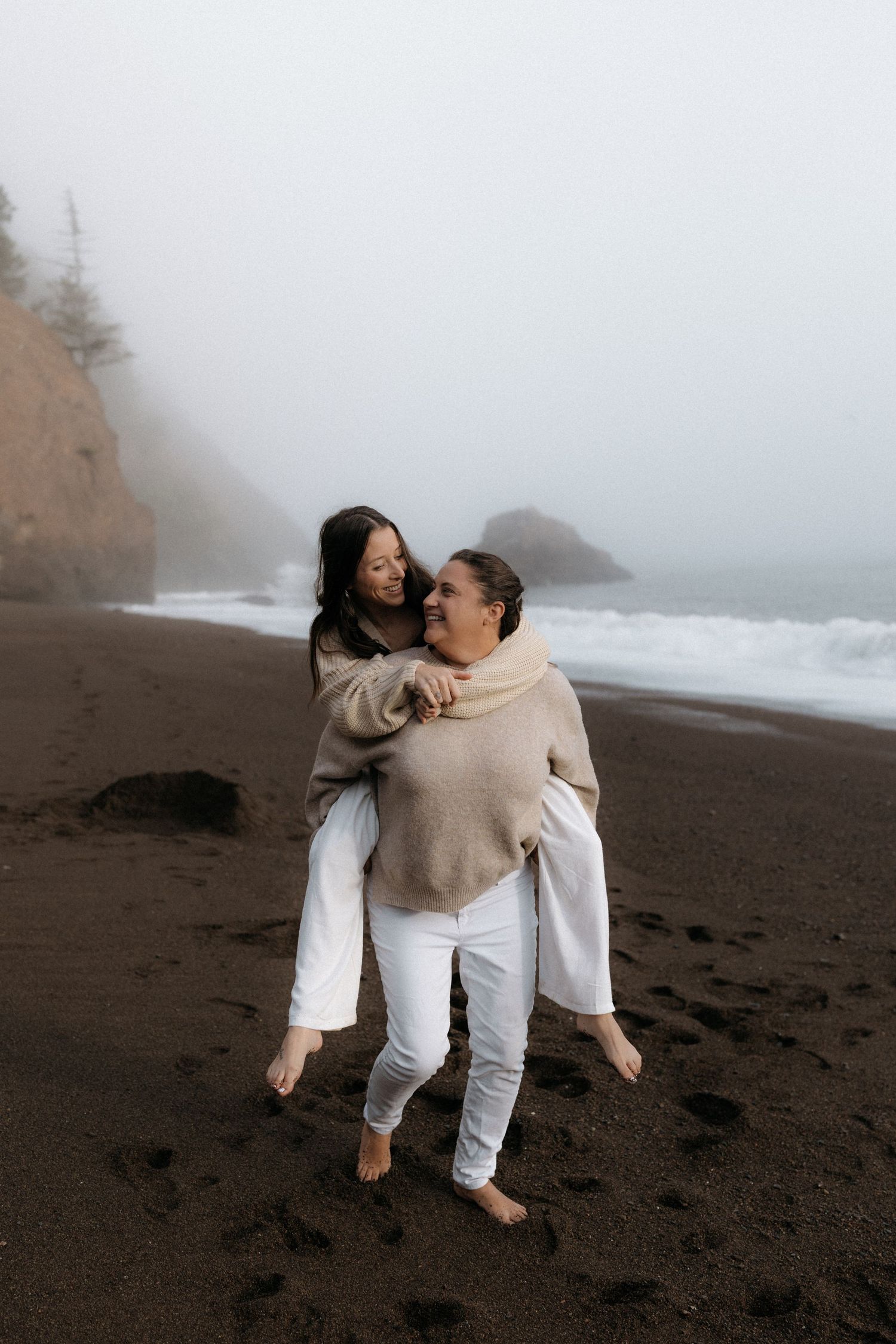 A couple in coordinated beige and white outfits sharing tender moments on a misty black sand beach.