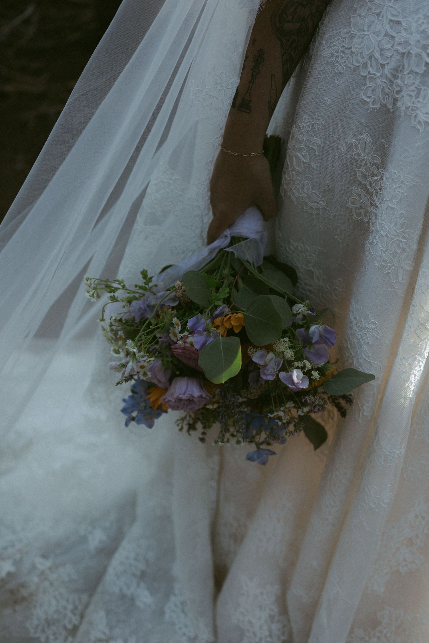 Bride in lace wedding dress holding a wildflower bouquet with purple, blue, and orange blooms, draped in a sheer veil.
