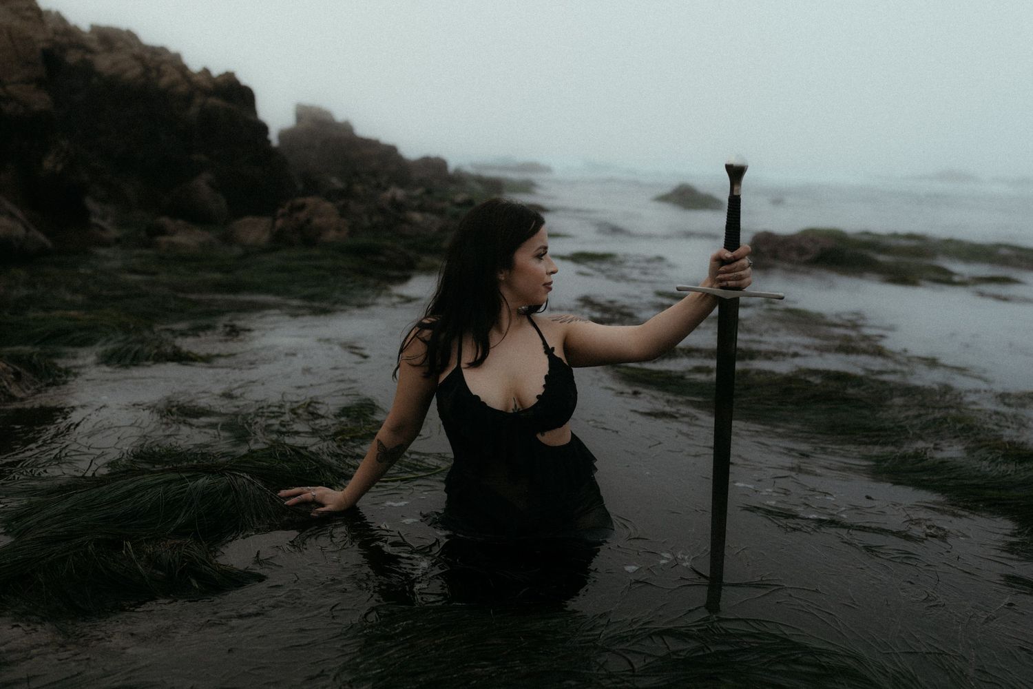 Figure in black poses dramatically on rocky coastal shore at dusk.