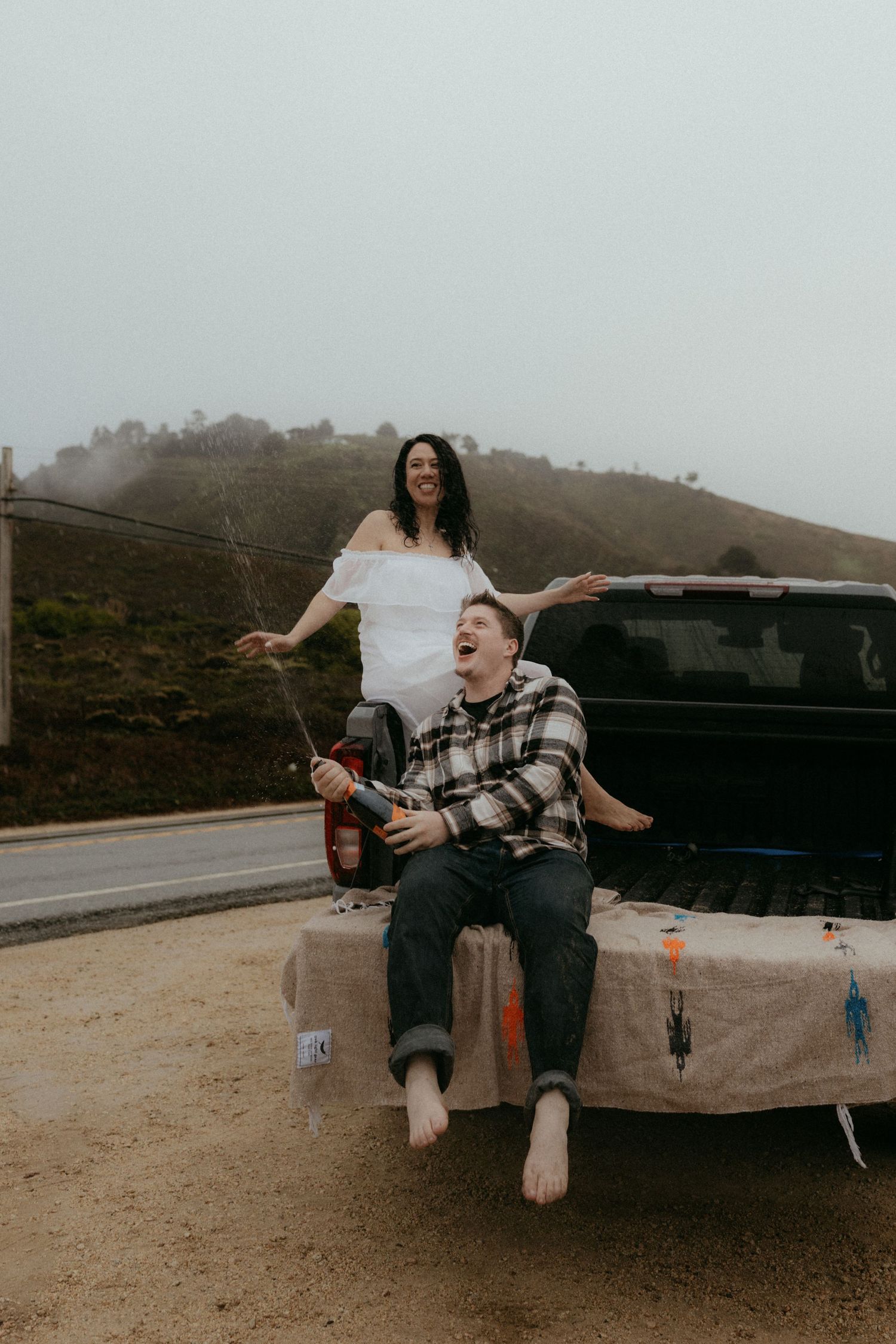 Playful couple laughing while sitting in pickup truck bed on coastal road.