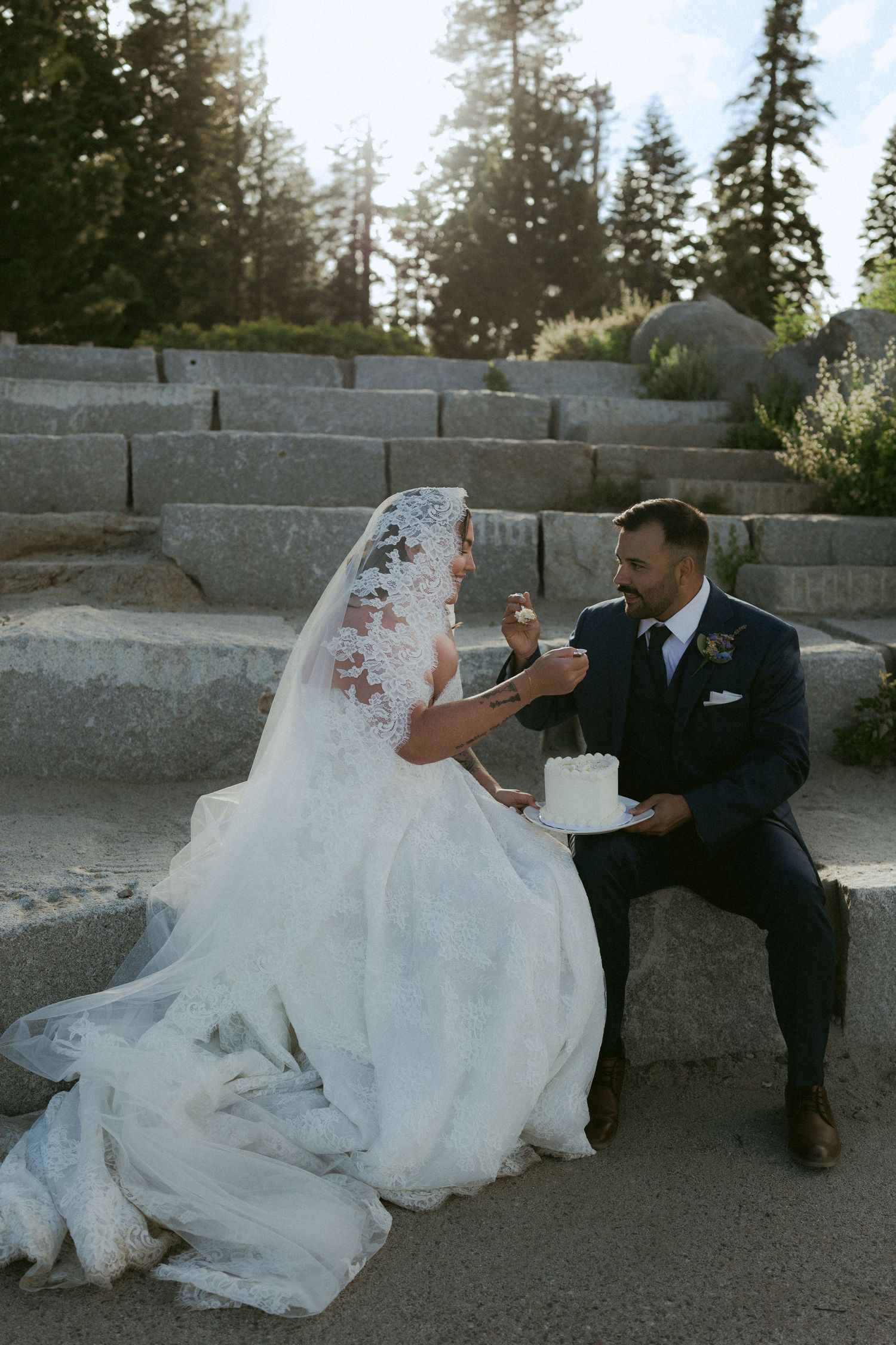 Bride and groom share wedding cake on stone steps surrounded by pine trees during outdoor elopement ceremony.