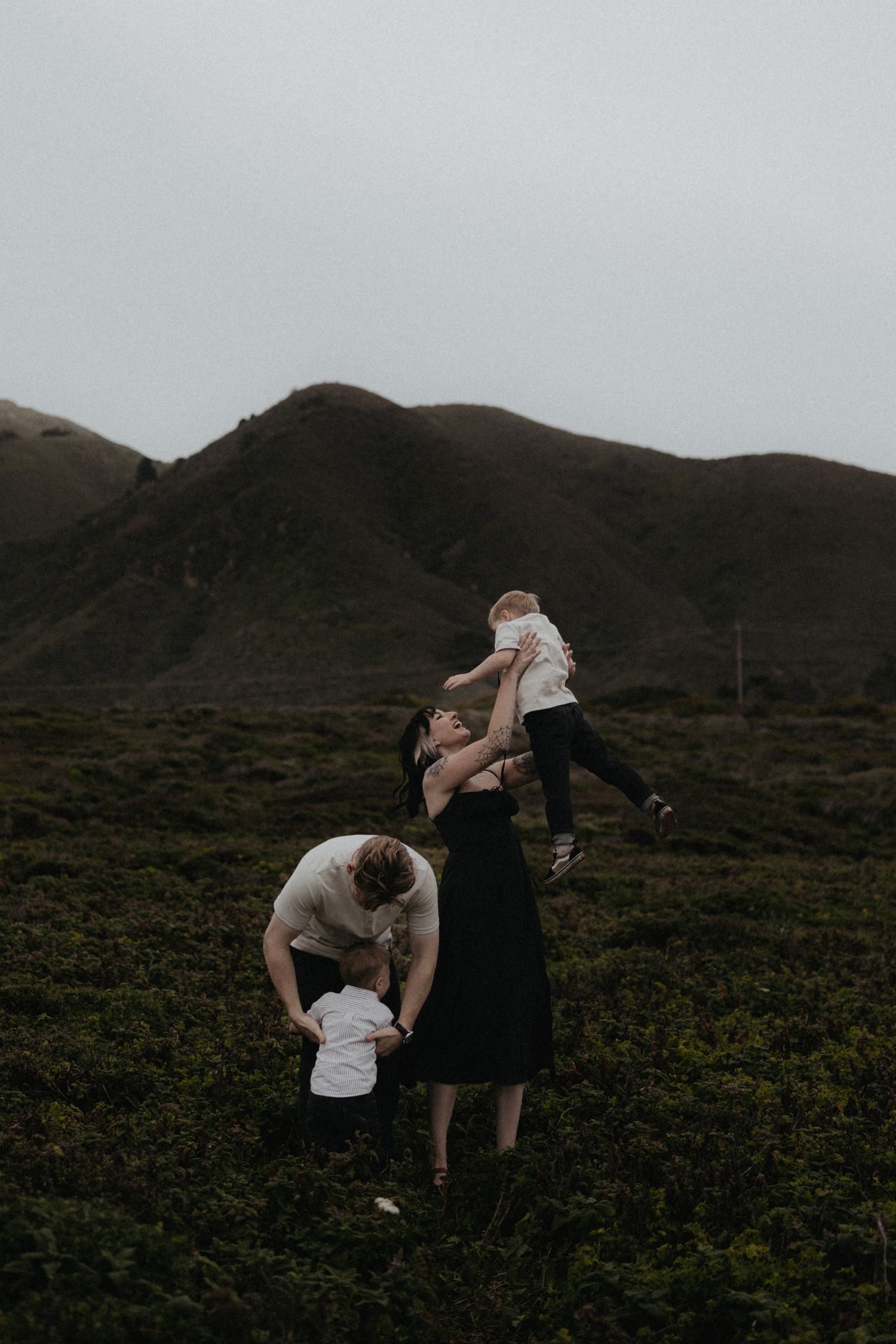 A family spends time together on a grassy hillside against a backdrop of mountains at dusk.