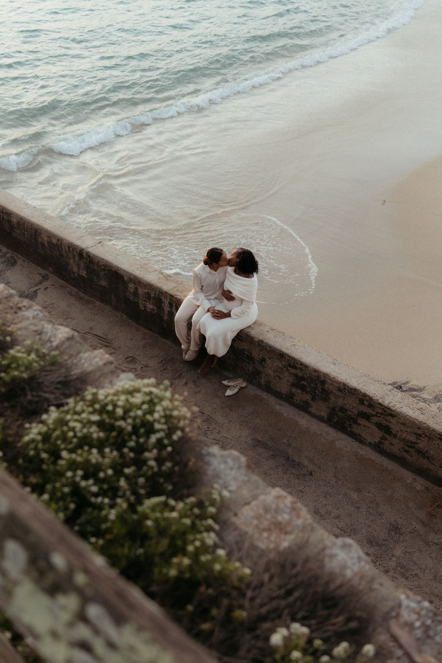 A couple sits intimately together on a stone wall overlooking the ocean waves at sunset.