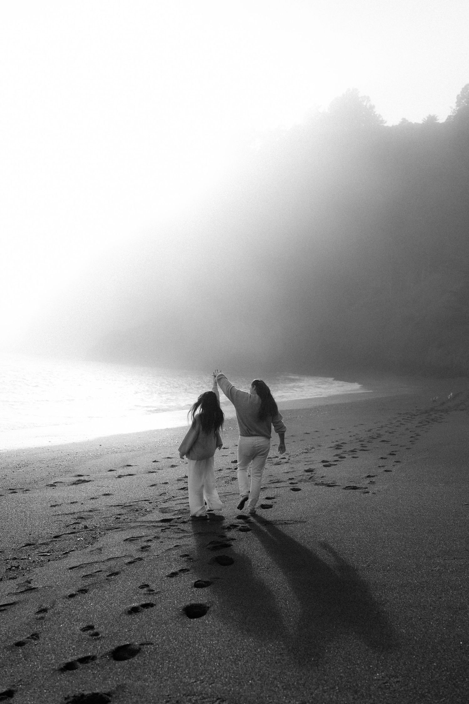 Two people walking hand in hand on a misty black sand beach leaving footprints in their wake.