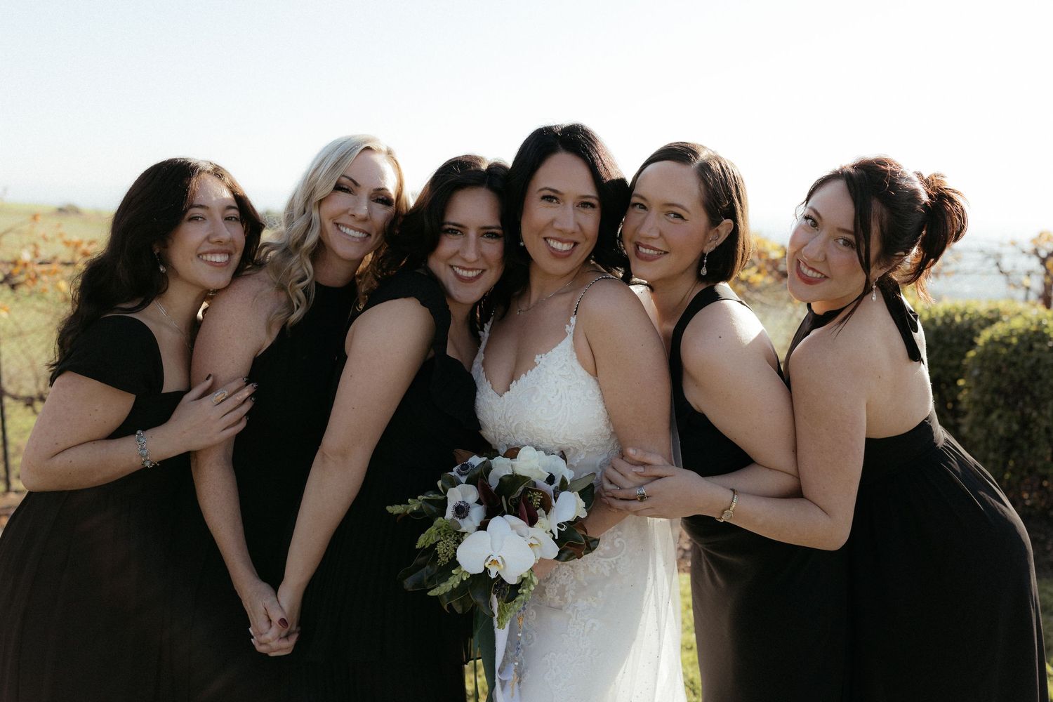 A group of women in black and white dresses pose together outdoors for wedding photos.