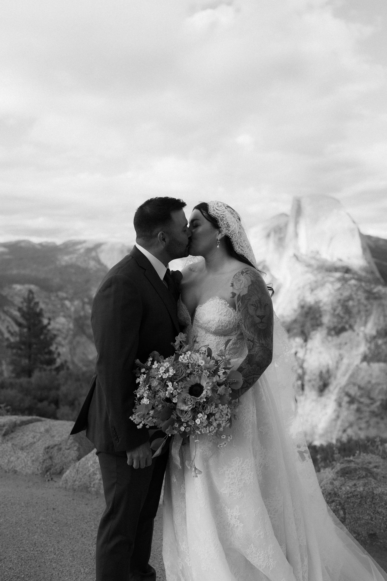 Bride and groom share a kiss at Yosemite with Half Dome in background, bride holding wildflower bouquet in black and white photo.