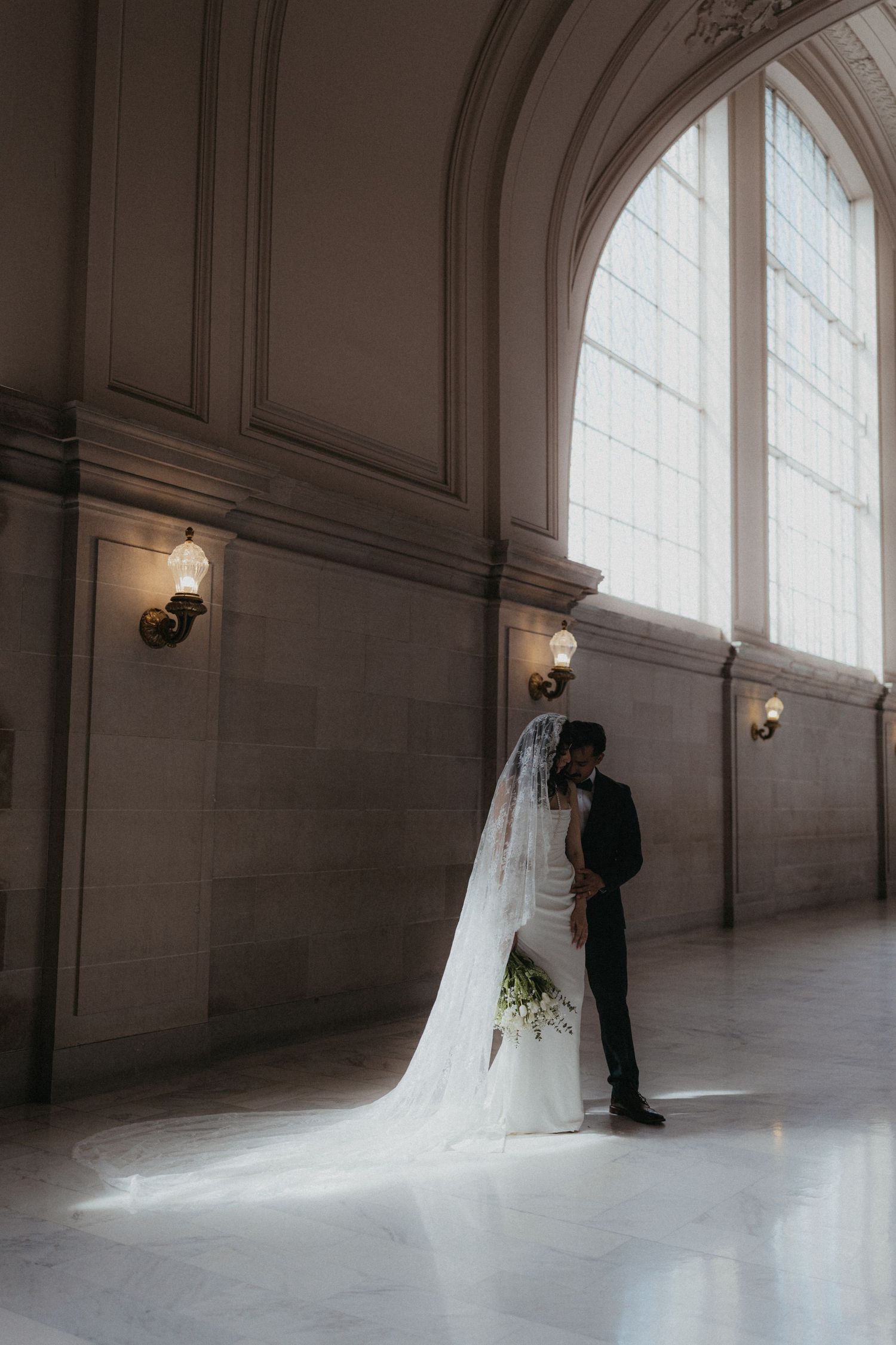 Bride in cathedral veil and groom share intimate moment by arched window at SF City Hall.