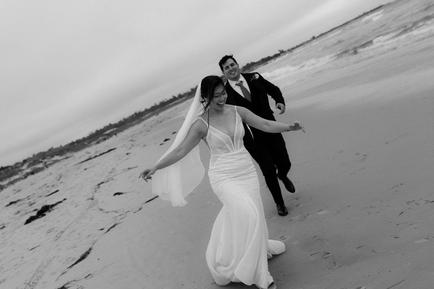 Black and white photo of a bride and groom laughing and running together on a sandy beach on their wedding day.