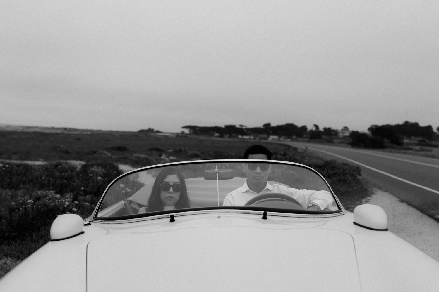 Black and white photo of two people riding in a classic white convertible car along a scenic coastal road.