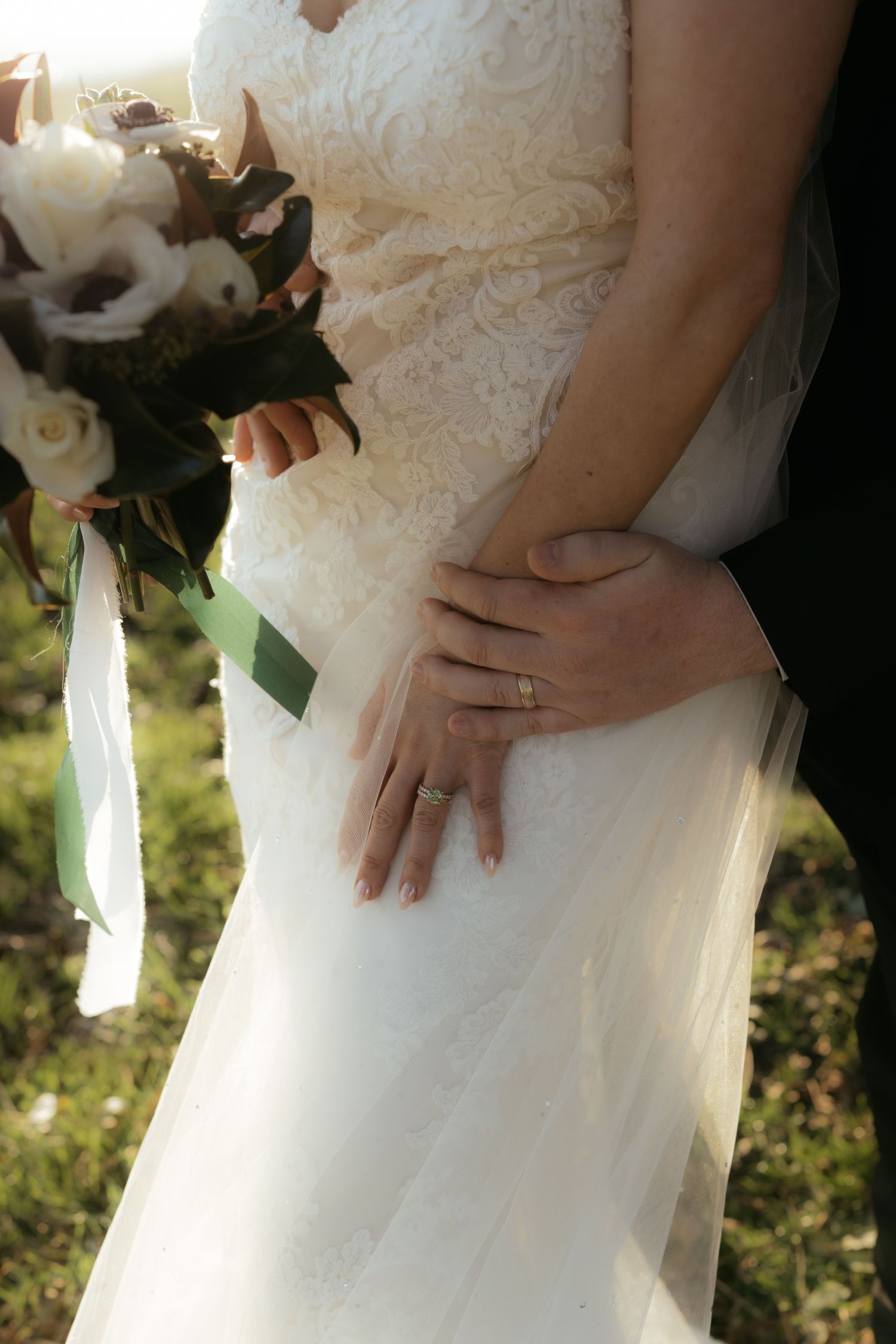 Close-up view of hands holding a white bridal bouquet against a lace wedding dress in natural sunlight.