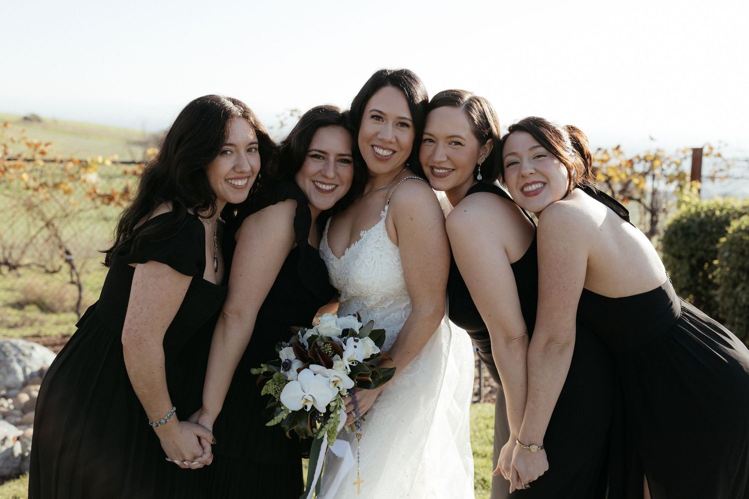A bridal party in black dresses and a white wedding gown pose together outdoors with joyful expressions.