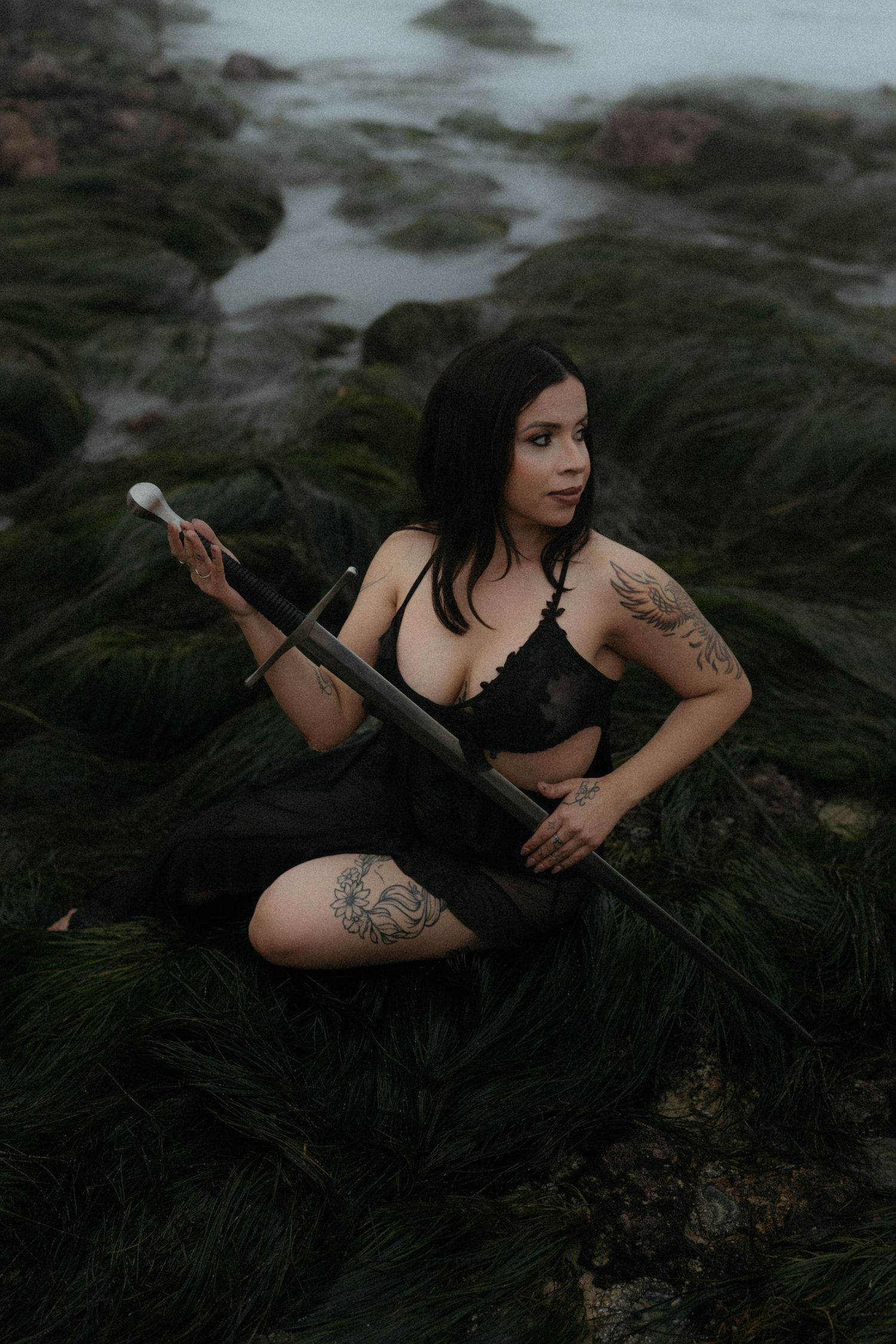 A figure in a black outfit poses artistically on dark rocks near water in a moody coastal setting.