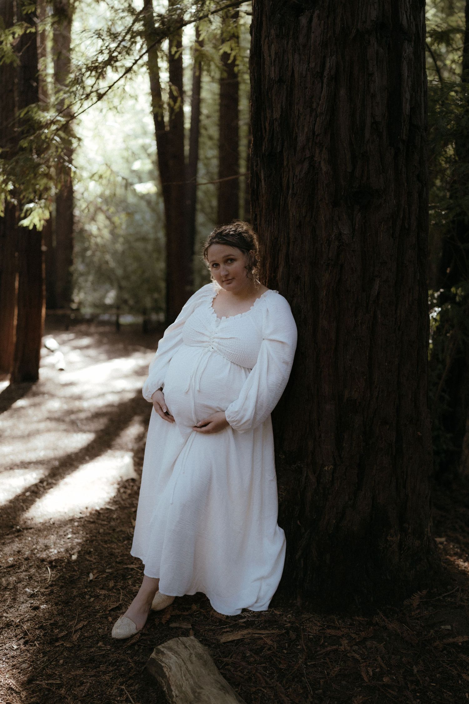 A person in a flowing white dress sits thoughtfully among tall redwood trees in a serene forest setting.
