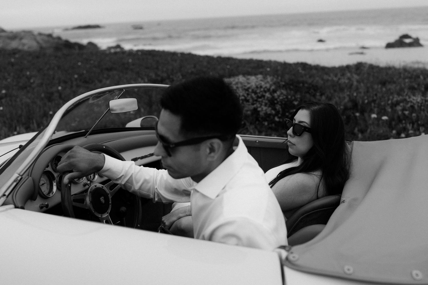Couple in a convertible car by the ocean, black and white photo, man driving, woman in sunglasses posing.