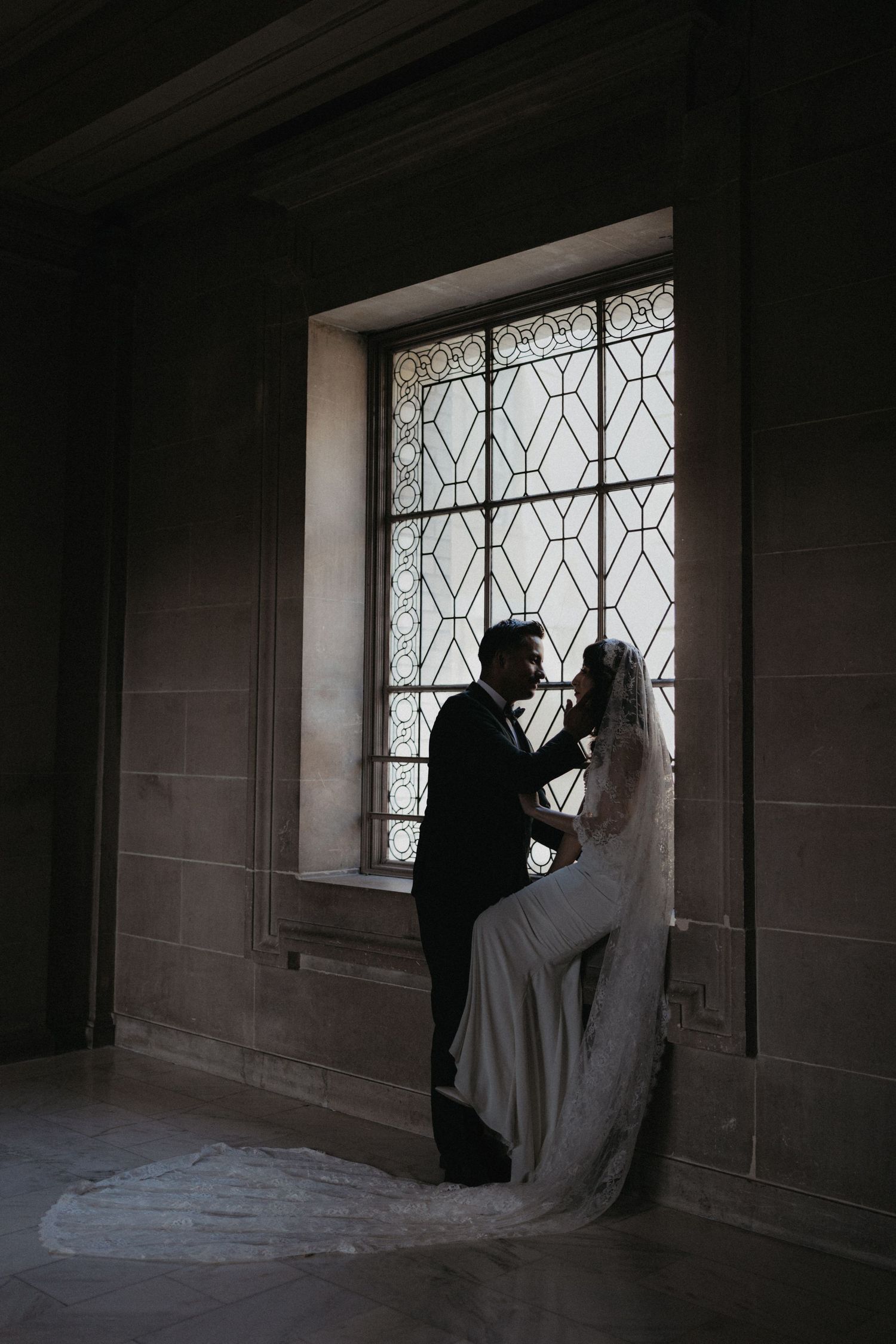 Bride and groom silhouetted against ornate stained glass window in dark stone building during romantic wedding portrait.