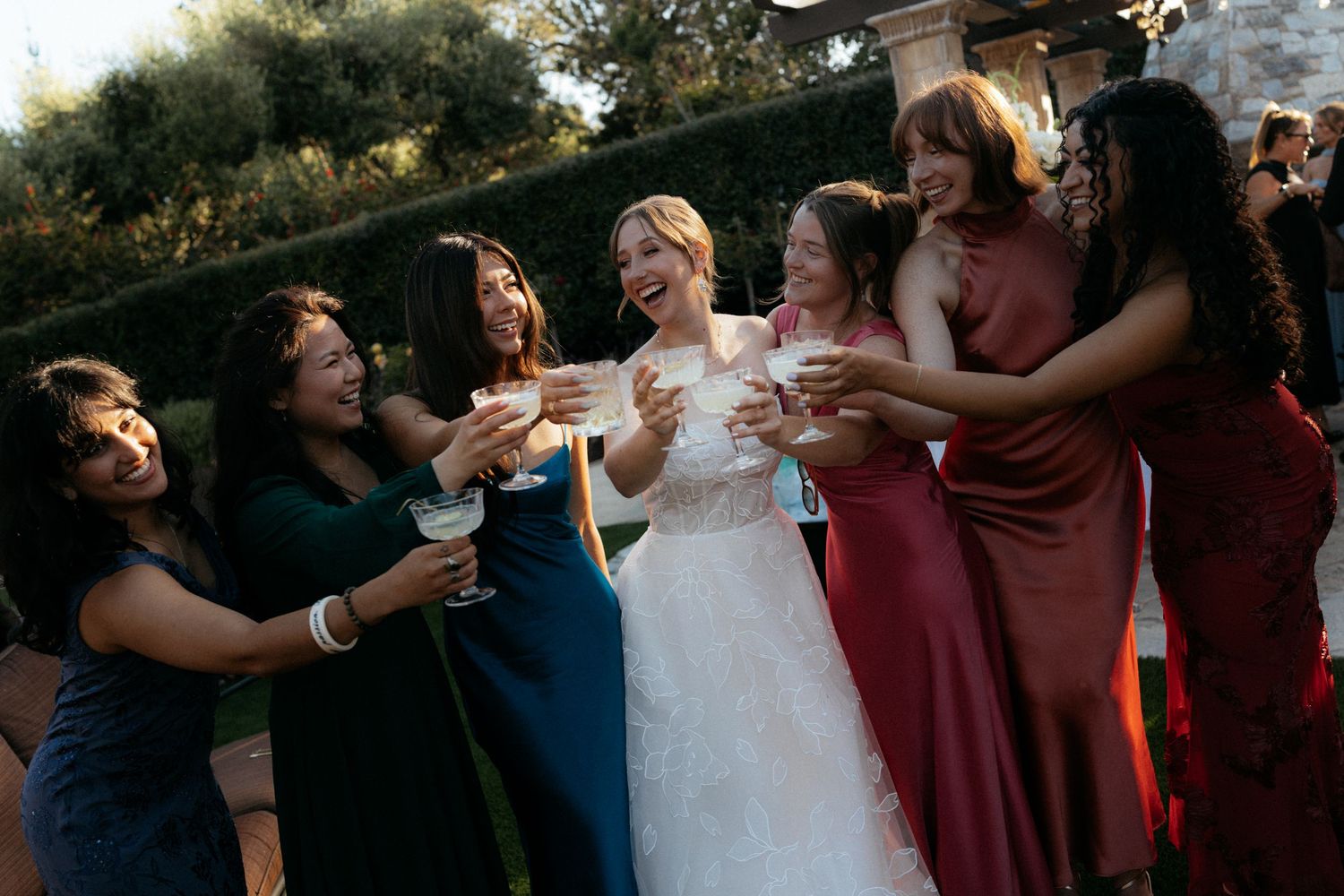 Bride in white gown toasting champagne with bridesmaids in colorful dresses at outdoor wedding celebration.