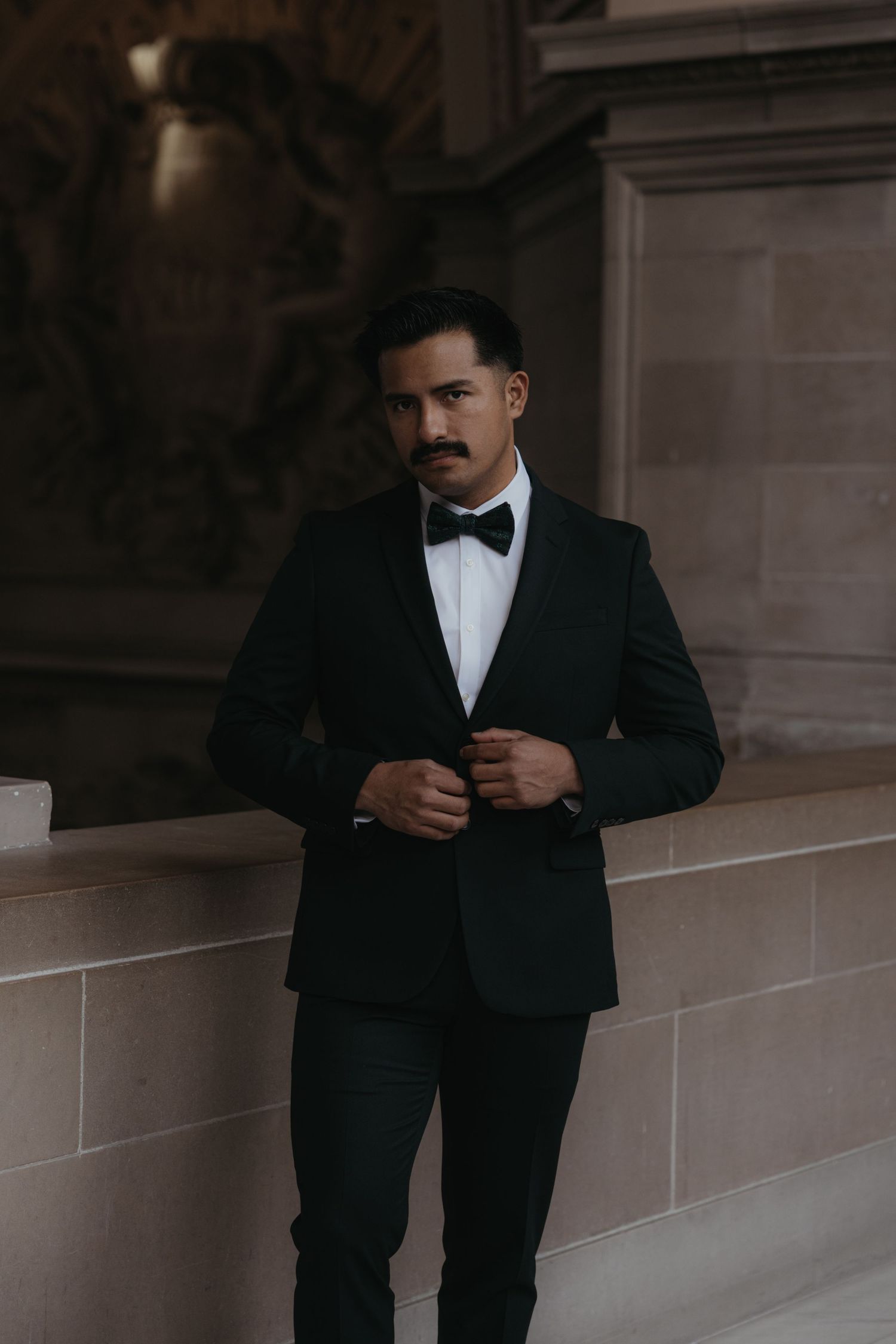 A groom in a classic black tuxedo and bow tie poses in an elegant architectural setting.