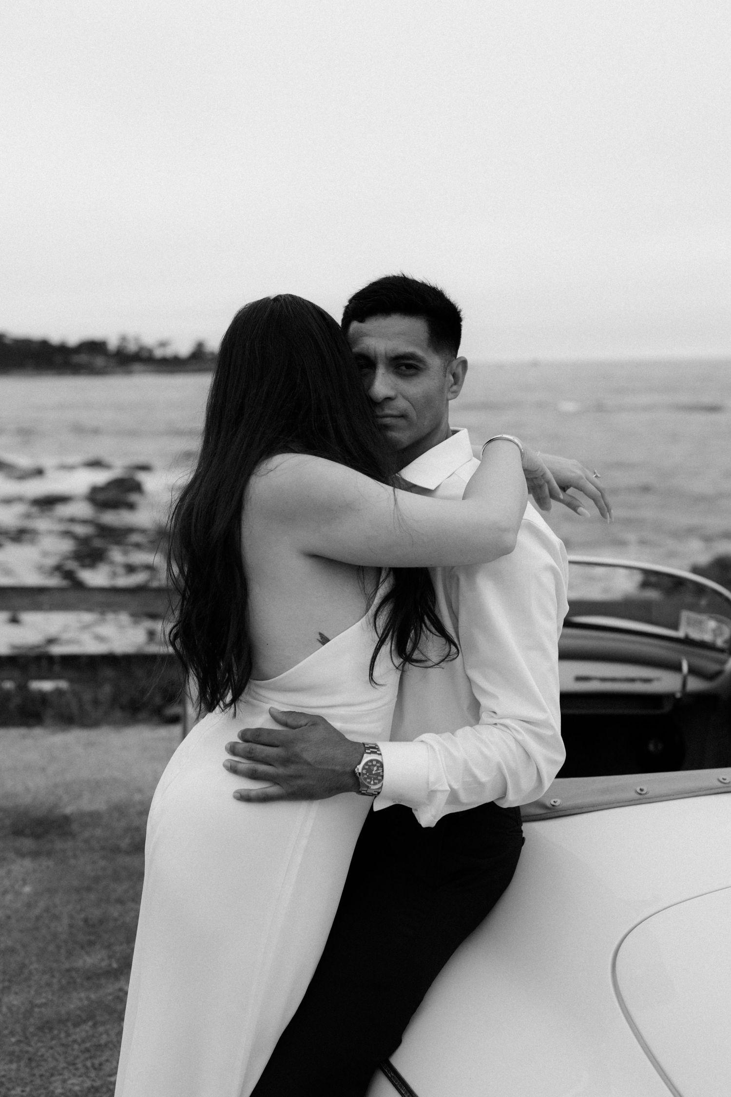 Couple embracing beside a vintage white convertible car near the ocean in a romantic black and white portrait.