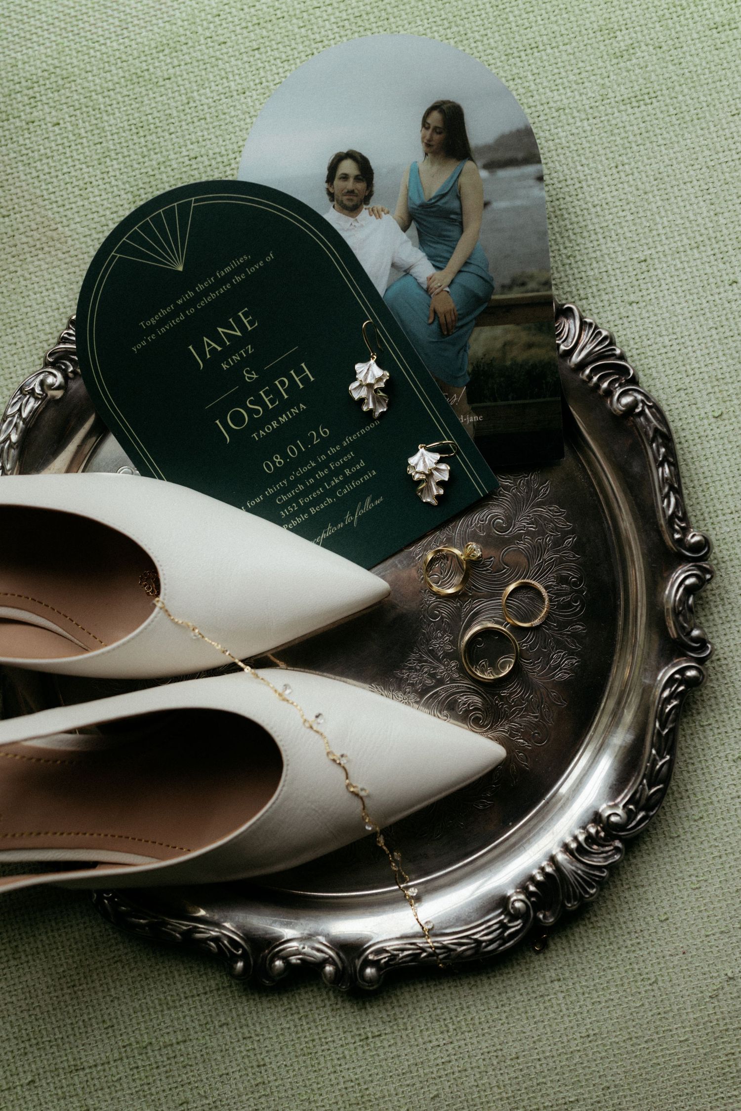 Wedding flat lay with white heels, gold rings, floral earrings, and elegant green arch invitation on silver tray.