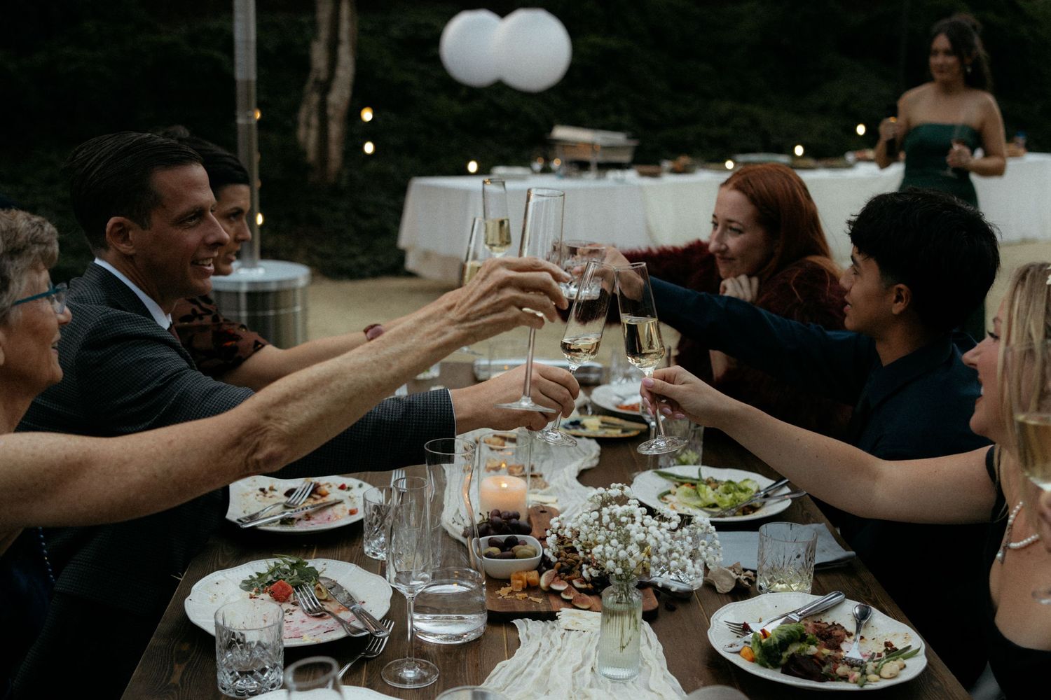 Guests raising wine glasses in a toast at an elegant outdoor dinner party with candlelit table settings.