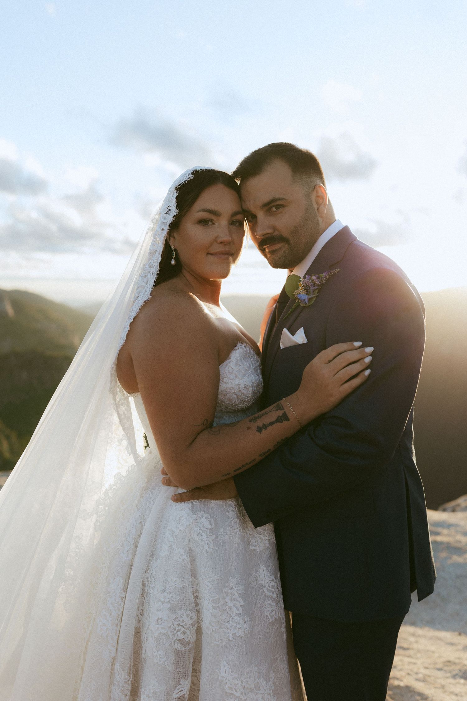 Bride and groom embracing at golden hour on mountain overlook in wedding attire.