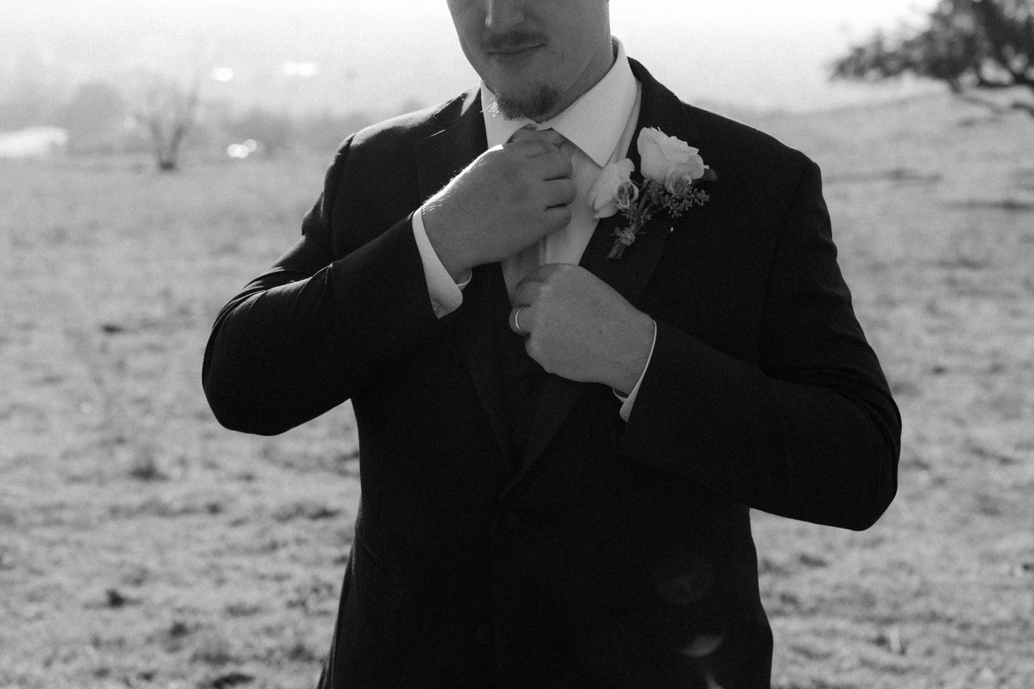 A groom in a black suit adjusts his tie while wearing a white boutonniere in a black and white photo.