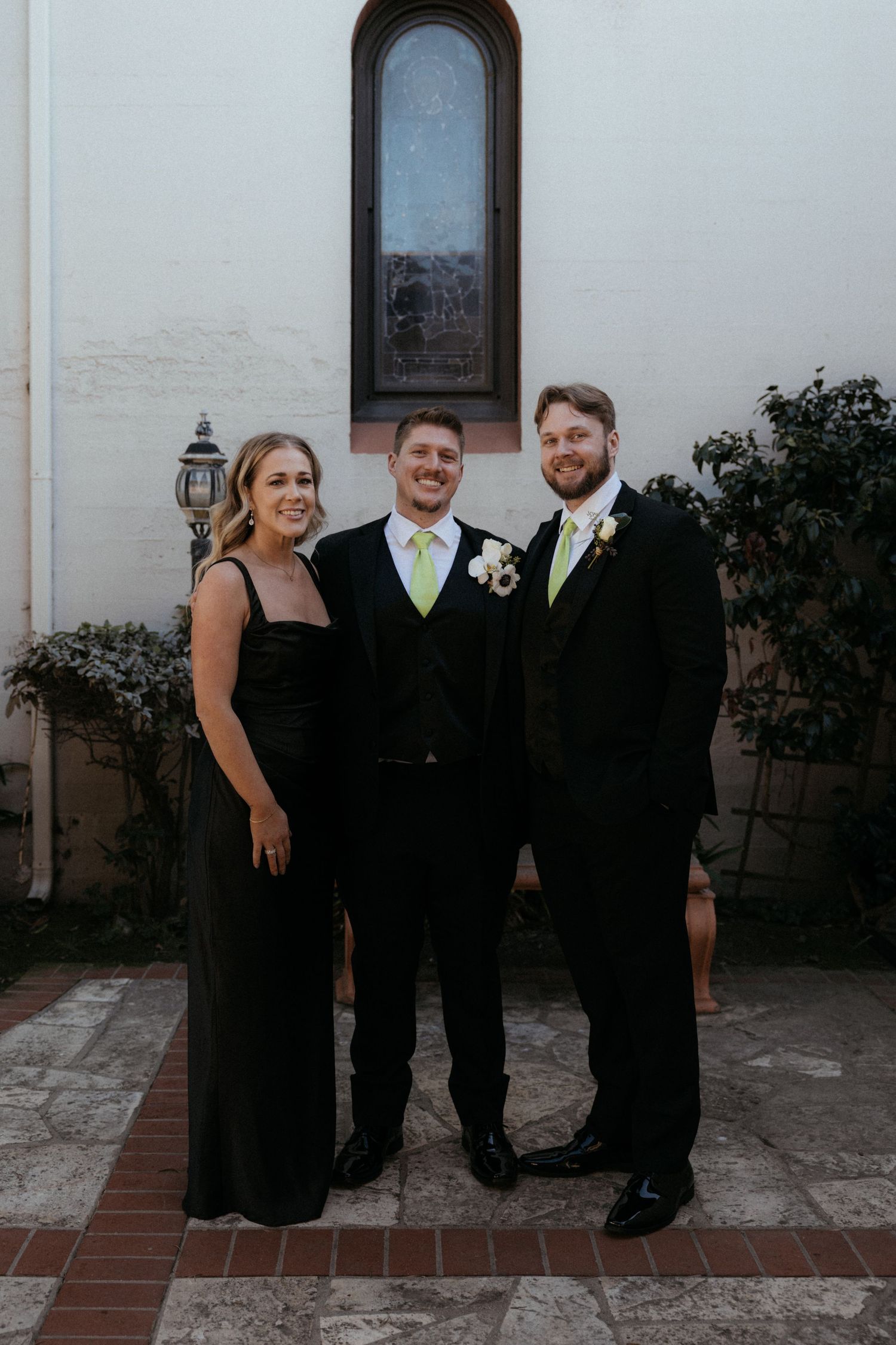 Wedding party members in black formal attire pose together outdoors against a white building wall at dusk.