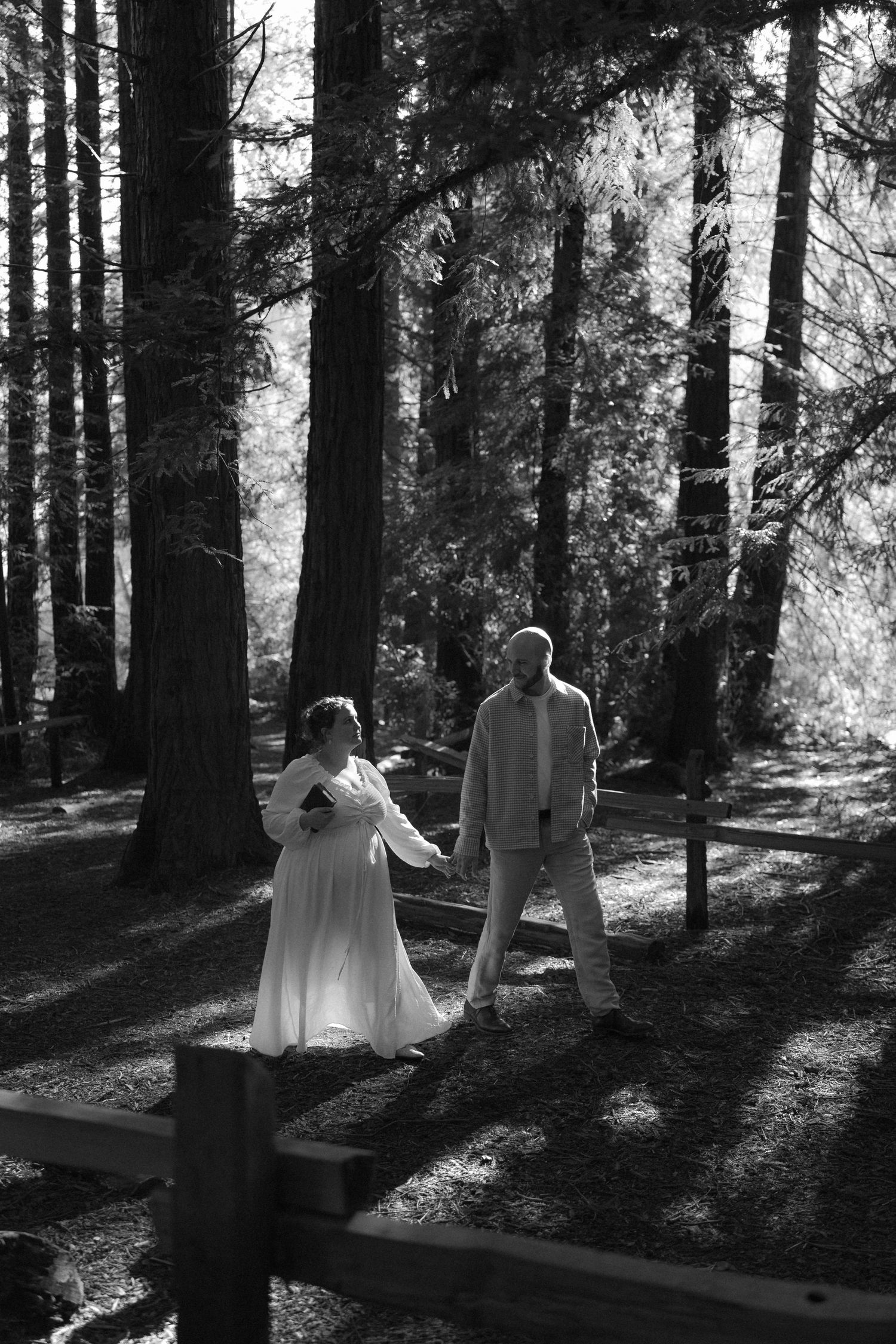 A romantic couple in white attire dances through sunlit redwood forest creating ethereal silhouettes.